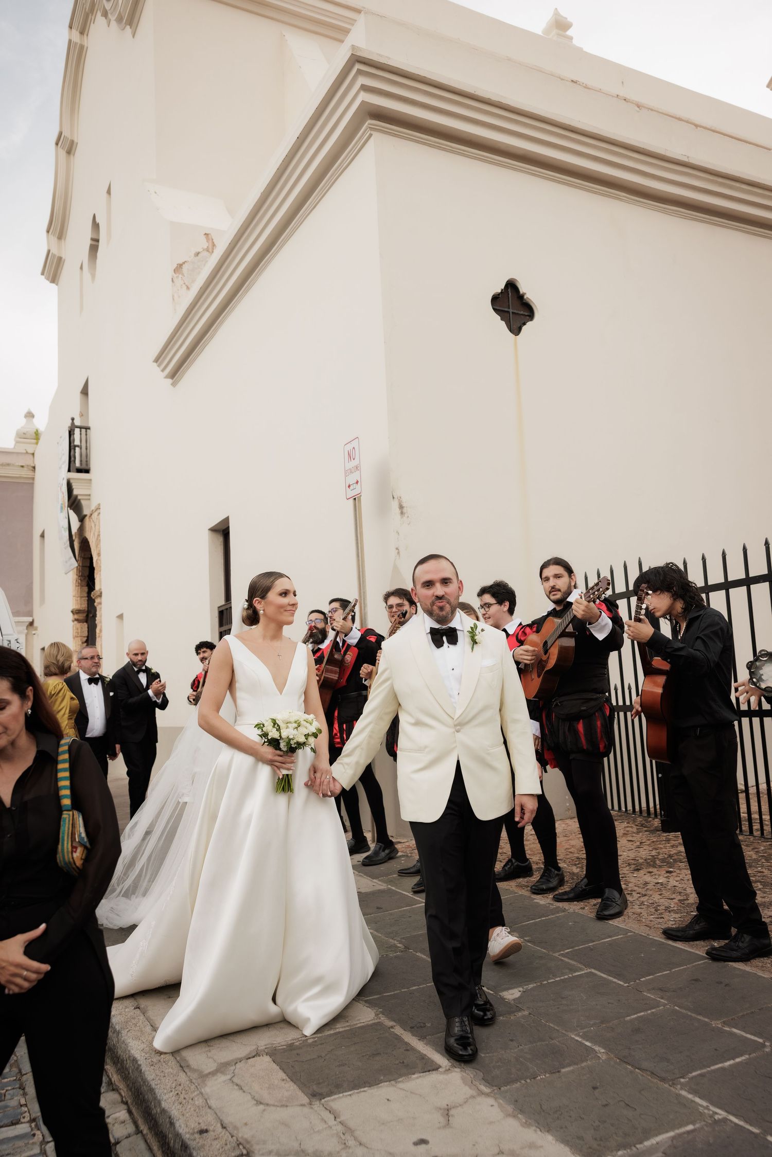 A couple exits a white church building while musicians with string instruments perform along the stone pathway.