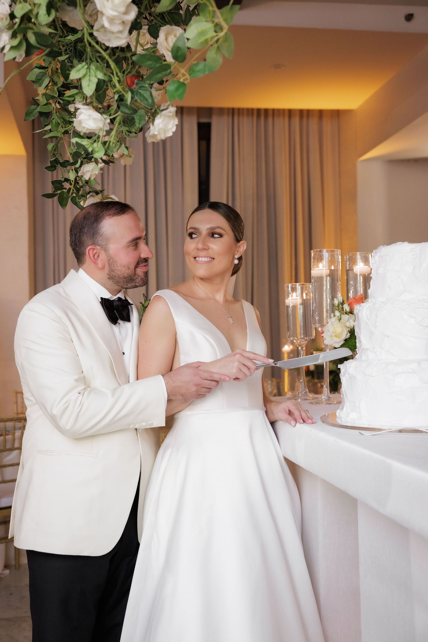 Newlyweds in white attire share a moment while cutting their elegant wedding cake adorned with floral decorations.