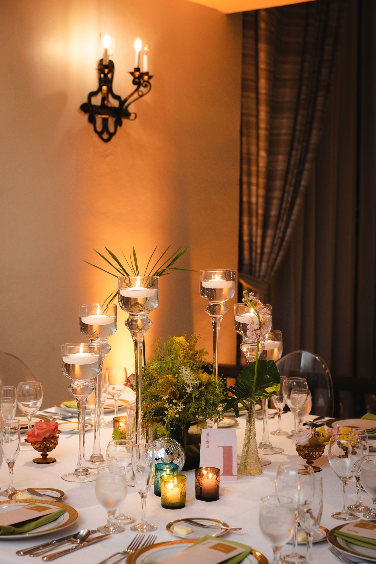 Elegant wedding reception table setting with glowing candles, glassware, and lush greenery centerpieces.