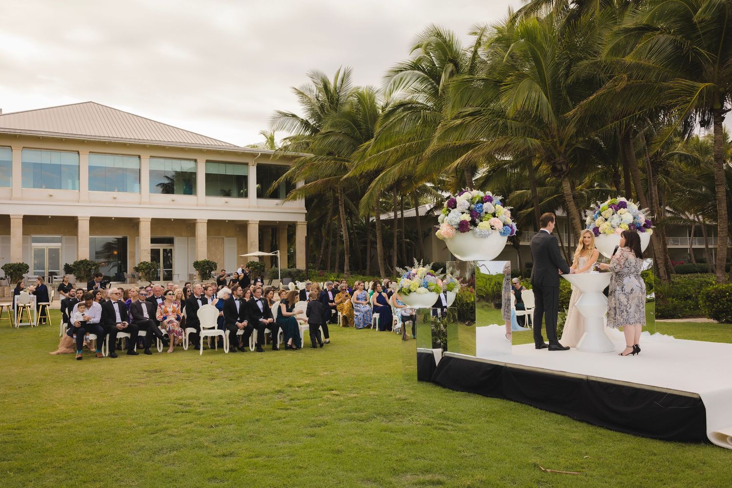 Outdoor wedding ceremony on a tropical lawn with palm trees and guests seated facing a raised platform stage.