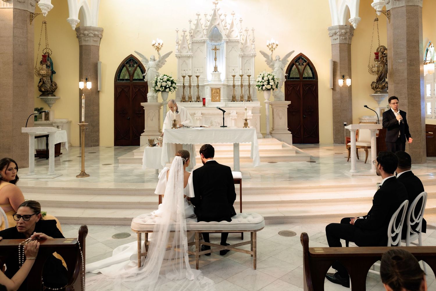 Traditional Catholic church wedding ceremony taking place in a white marble sanctuary with Gothic arches and altar decorations.
