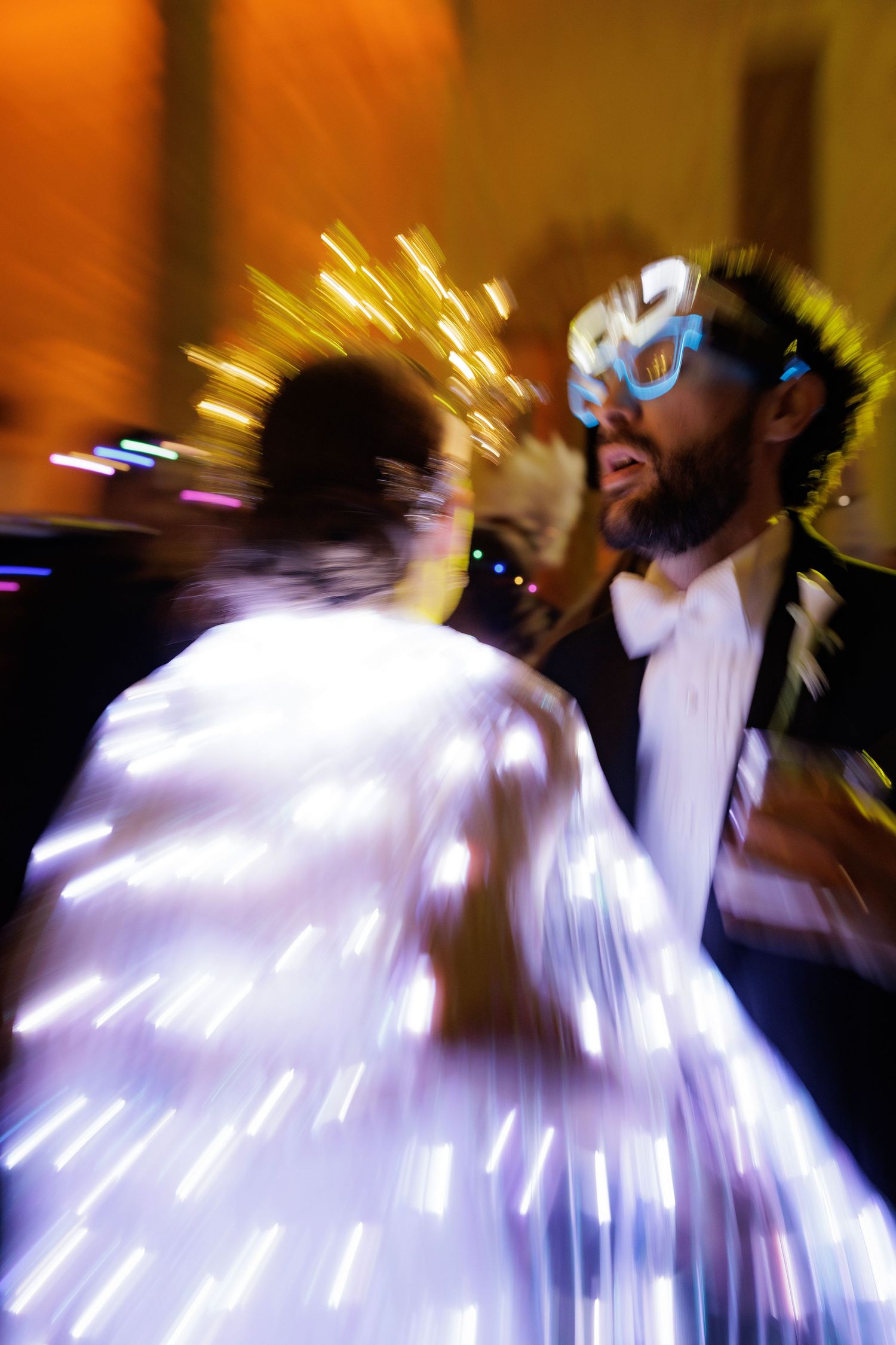 Motion-blurred sequence captures dancers in white dresses twirling with dramatic light trails against orange backdrop.