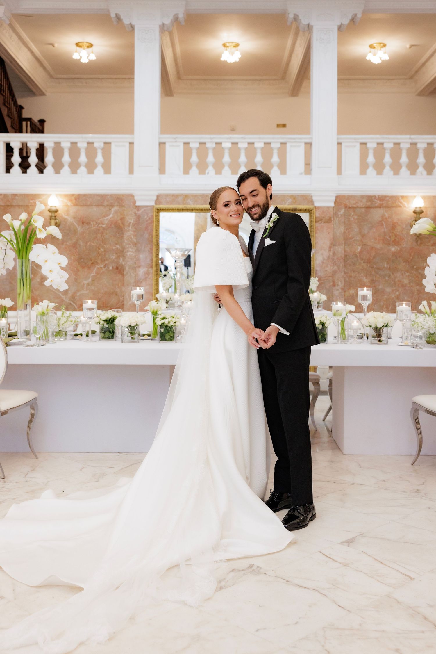 A couple in a luxurious white wedding venue featuring elegant columns, marble flooring and floral decorations.