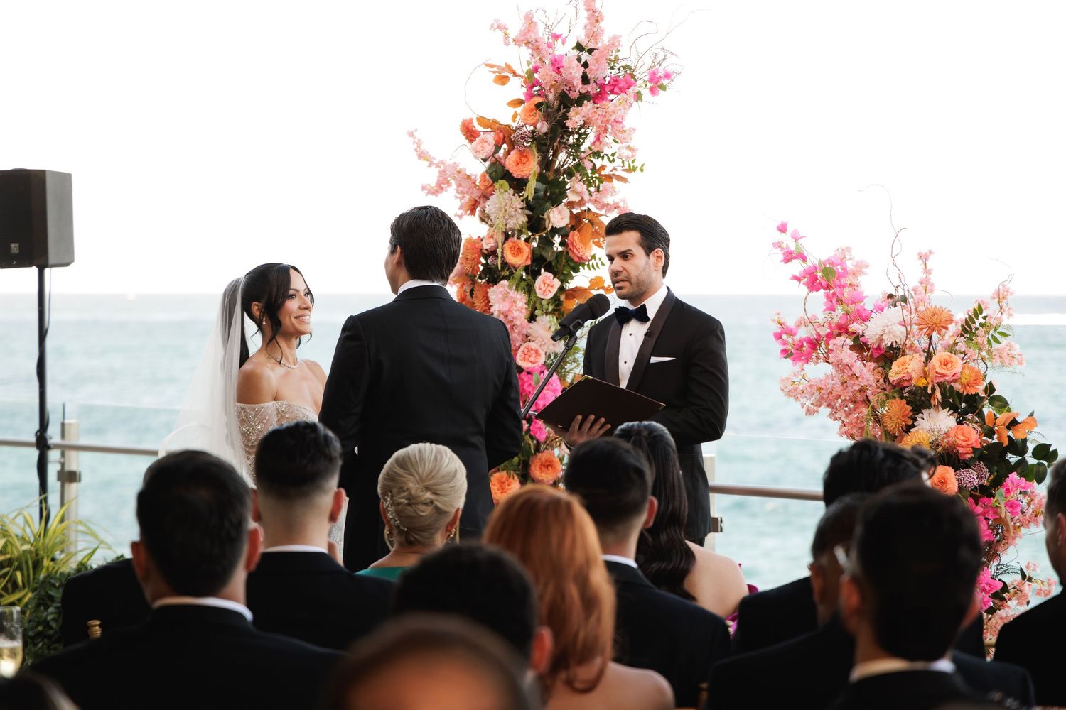 Wedding guests observe a waterfront ceremony with vibrant pink floral arrangements decorating the outdoor venue.