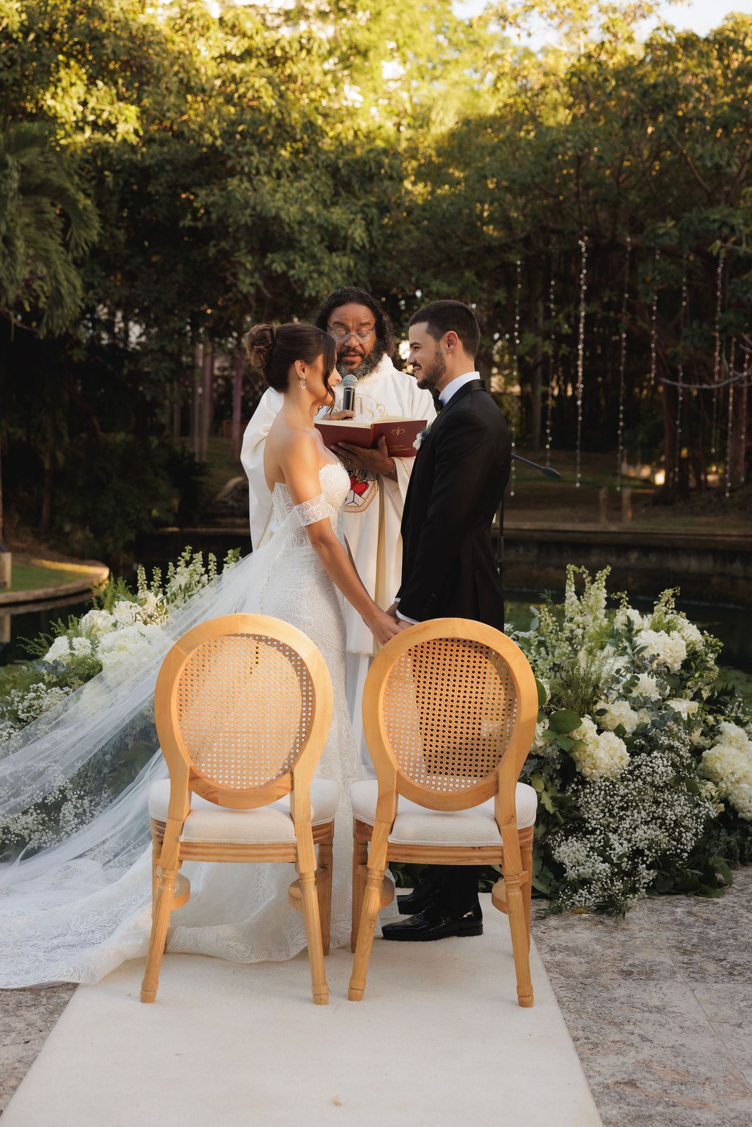 Outdoor wedding ceremony in a lush garden setting with elegant cane chairs and white floral arrangements at sunset.