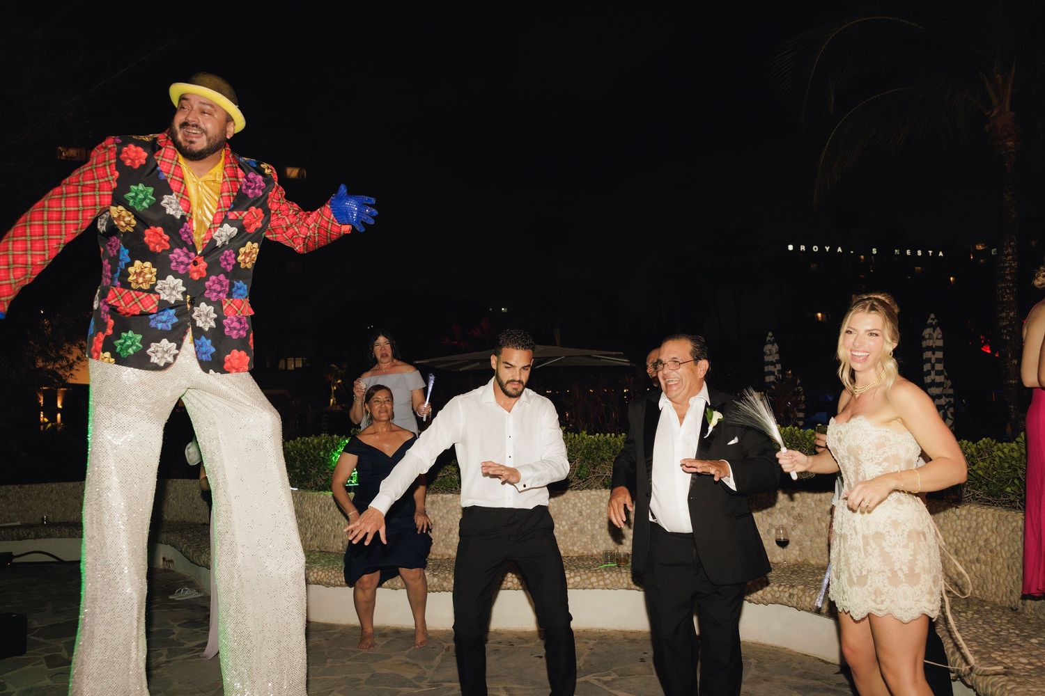 A stilt walker in a colorful floral shirt entertains party guests dancing in formal attire at an outdoor evening event.