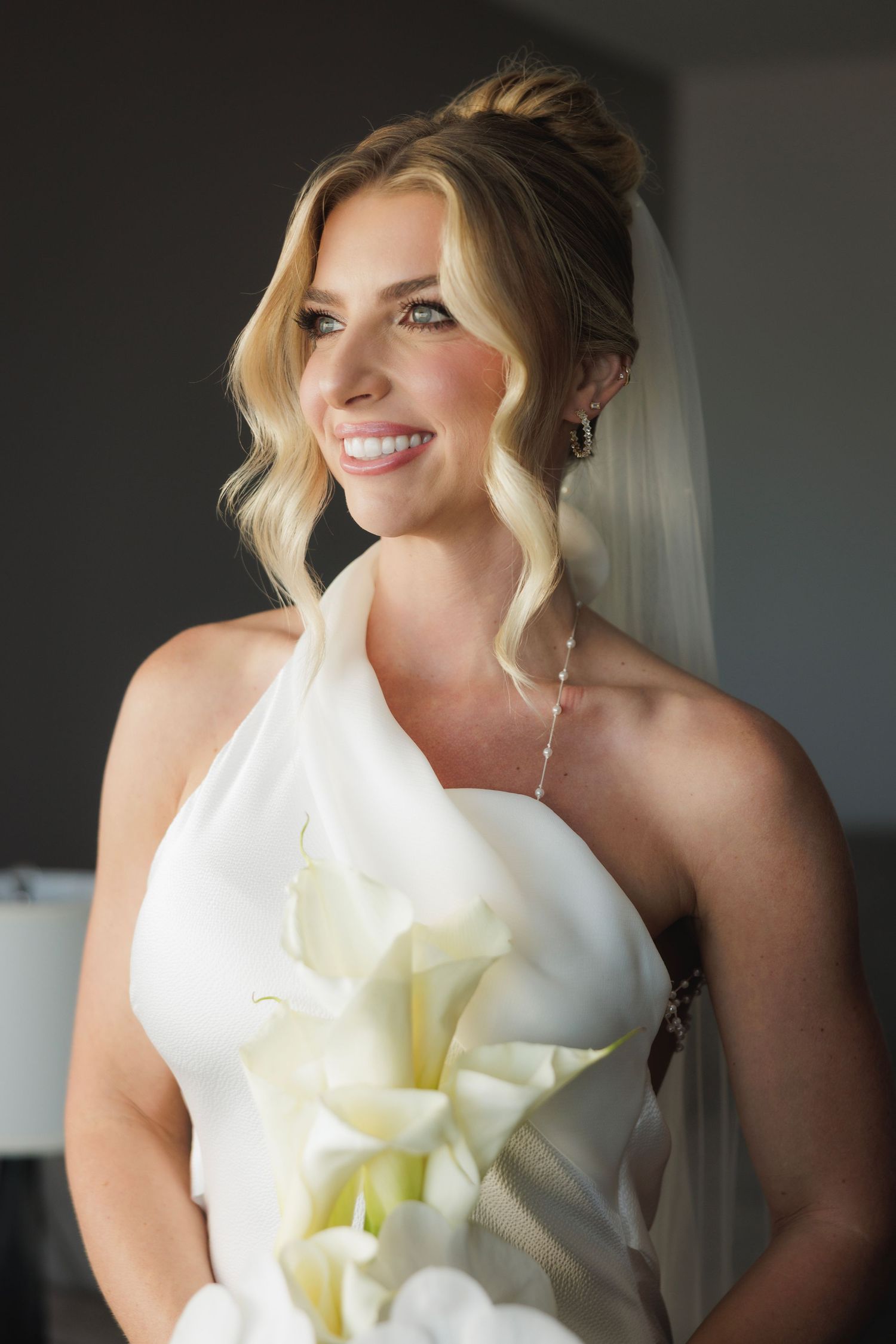Bride in elegant one-shoulder white wedding dress with cascading blonde waves and calla lily bouquet smiles in natural light.
