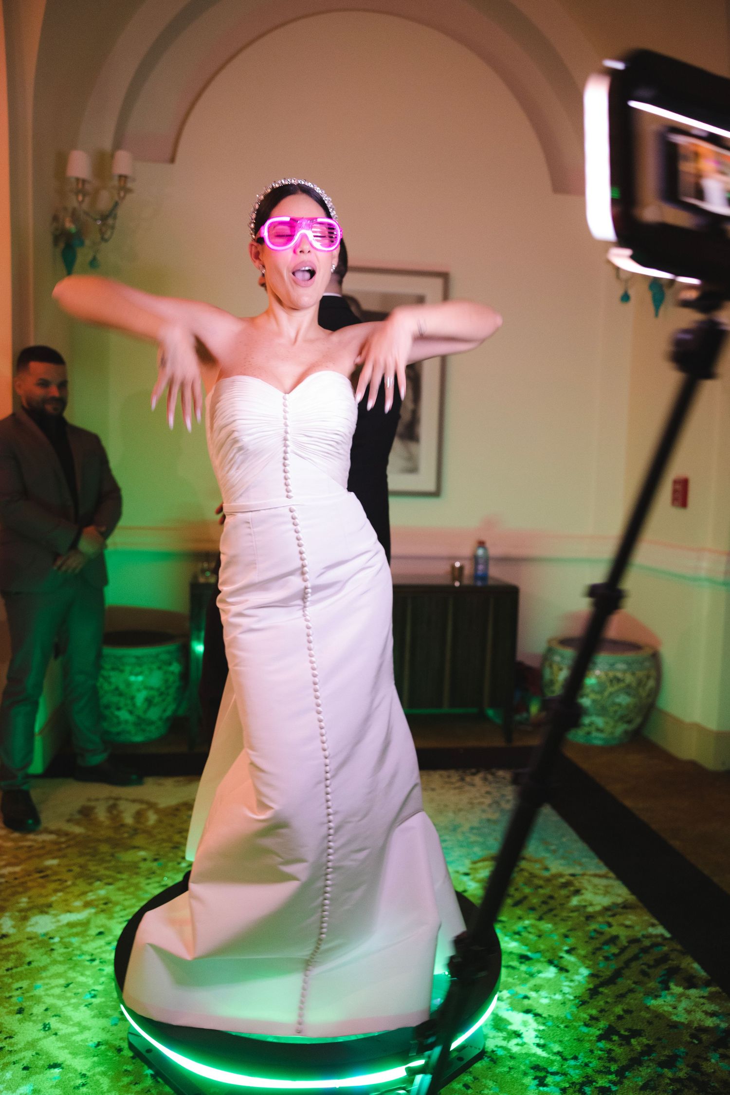 A bride wearing pink party glasses dances playfully on an illuminated platform at a wedding reception with green lighting.
