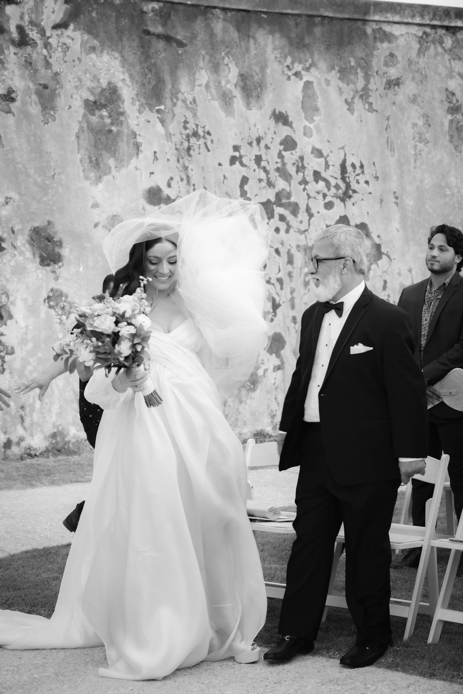 A wedding ceremony unfolds against a weathered stone wall backdrop with formal attire and bouquets.