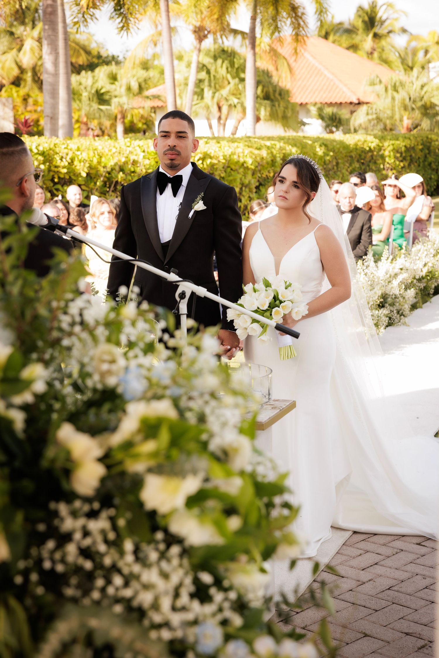 A tropical wedding ceremony with palm trees, white floral arrangements and a brick path in the background.