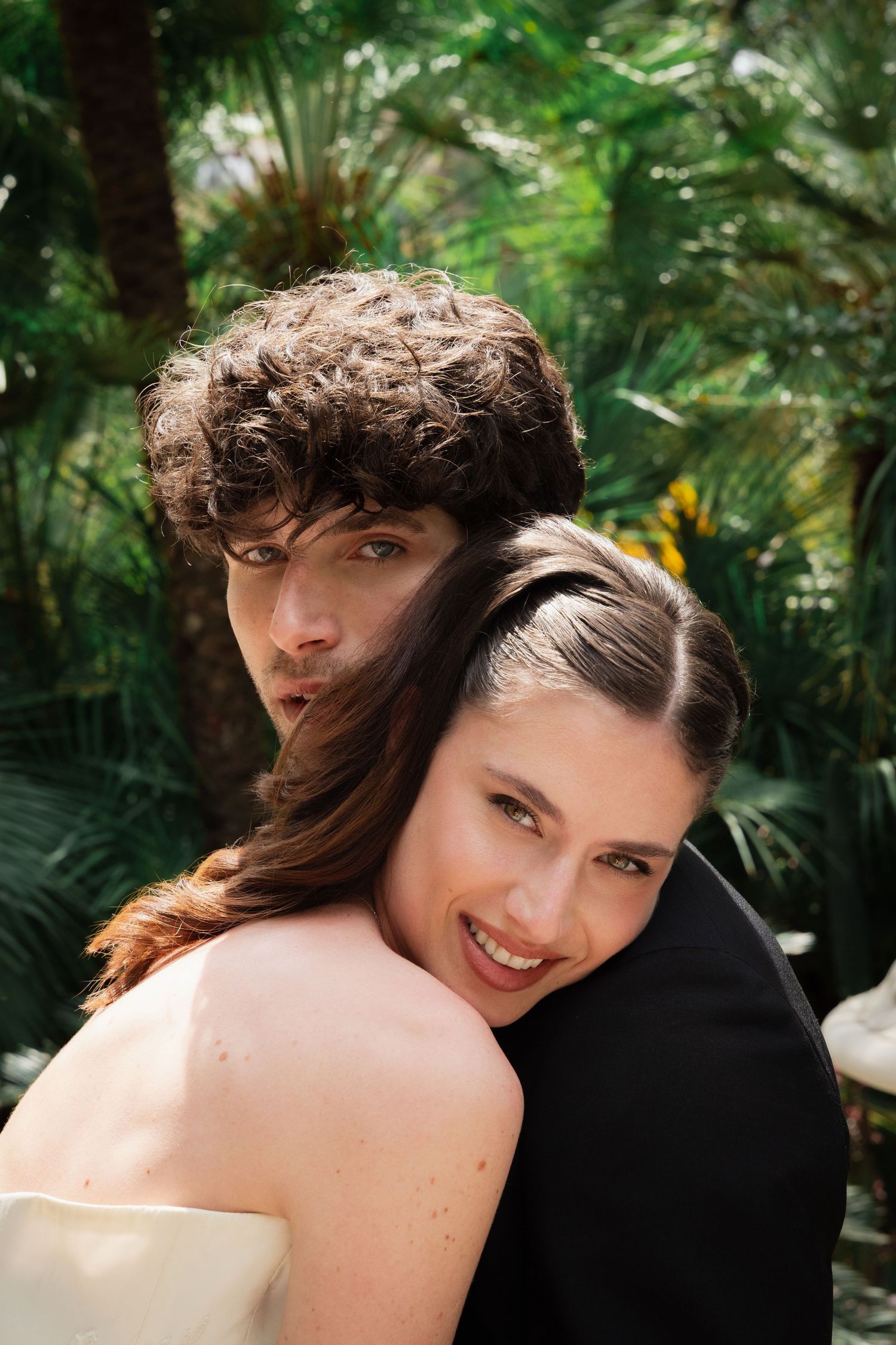A smiling bride in a strapless gown embraces a curly-haired groom in a tropical garden setting.