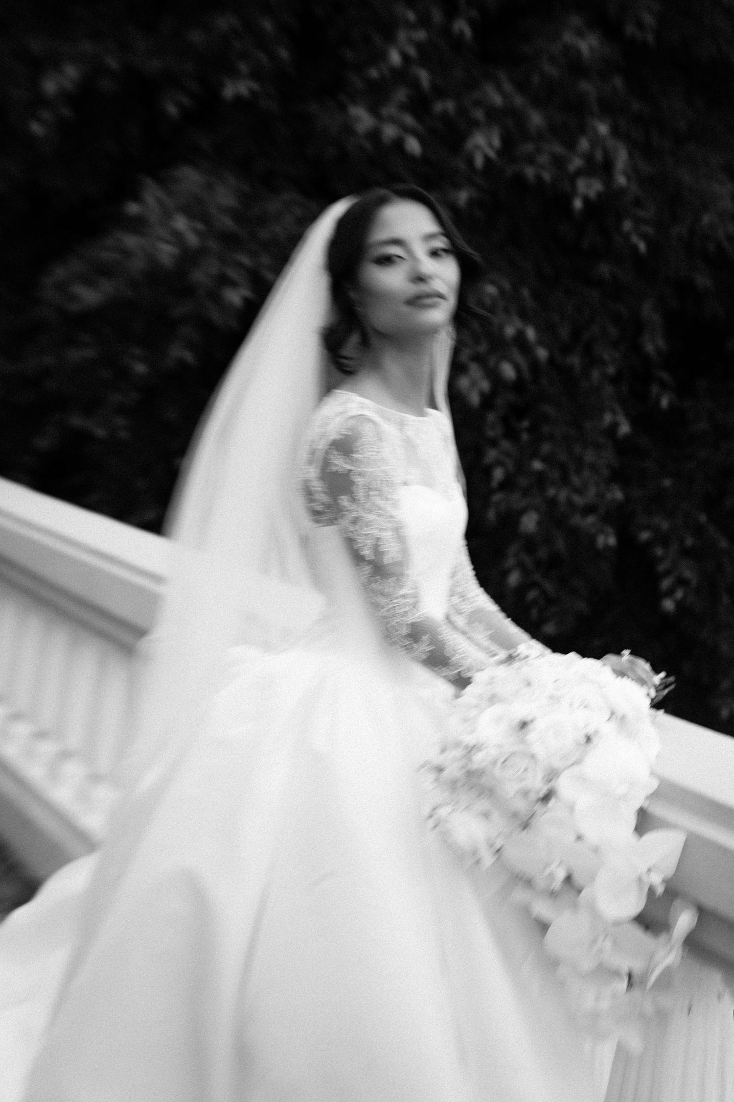 Black and white photo of a bride in a lace gown and veil holding a bouquet, posing gracefully on a white balcony.