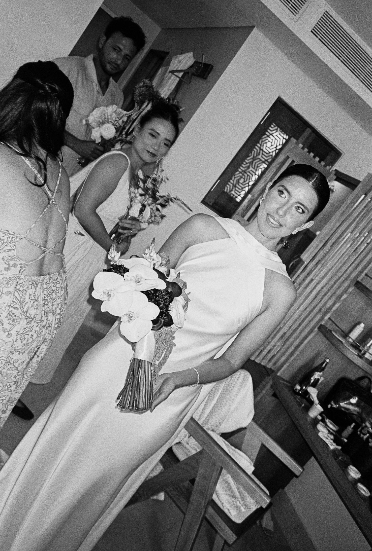 Smiling bride in white gown holds bouquet surrounded by bridesmaids in black and white wedding photo.