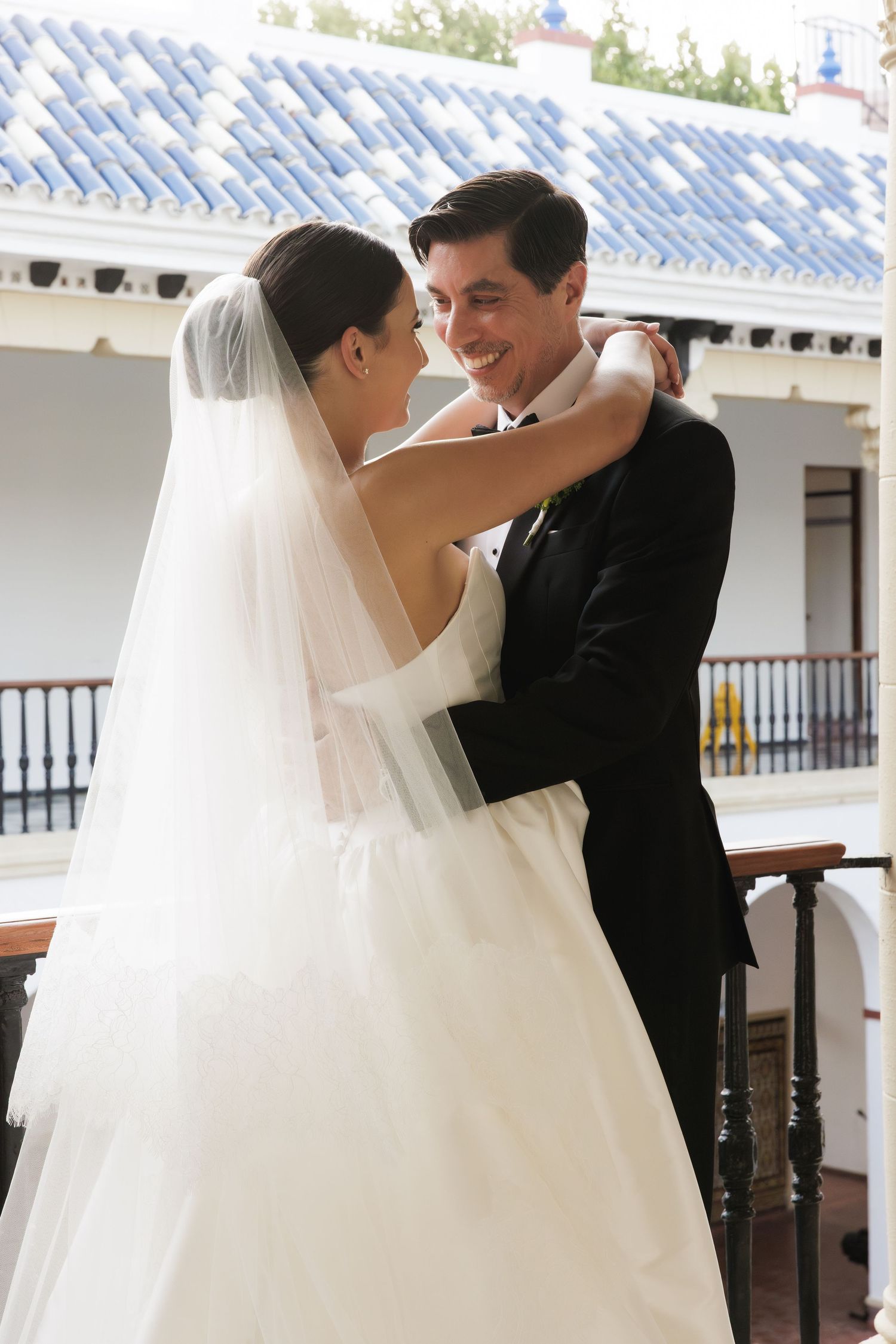 A joyful wedding couple embraces on a covered porch with white architectural details and blue tiles overhead.