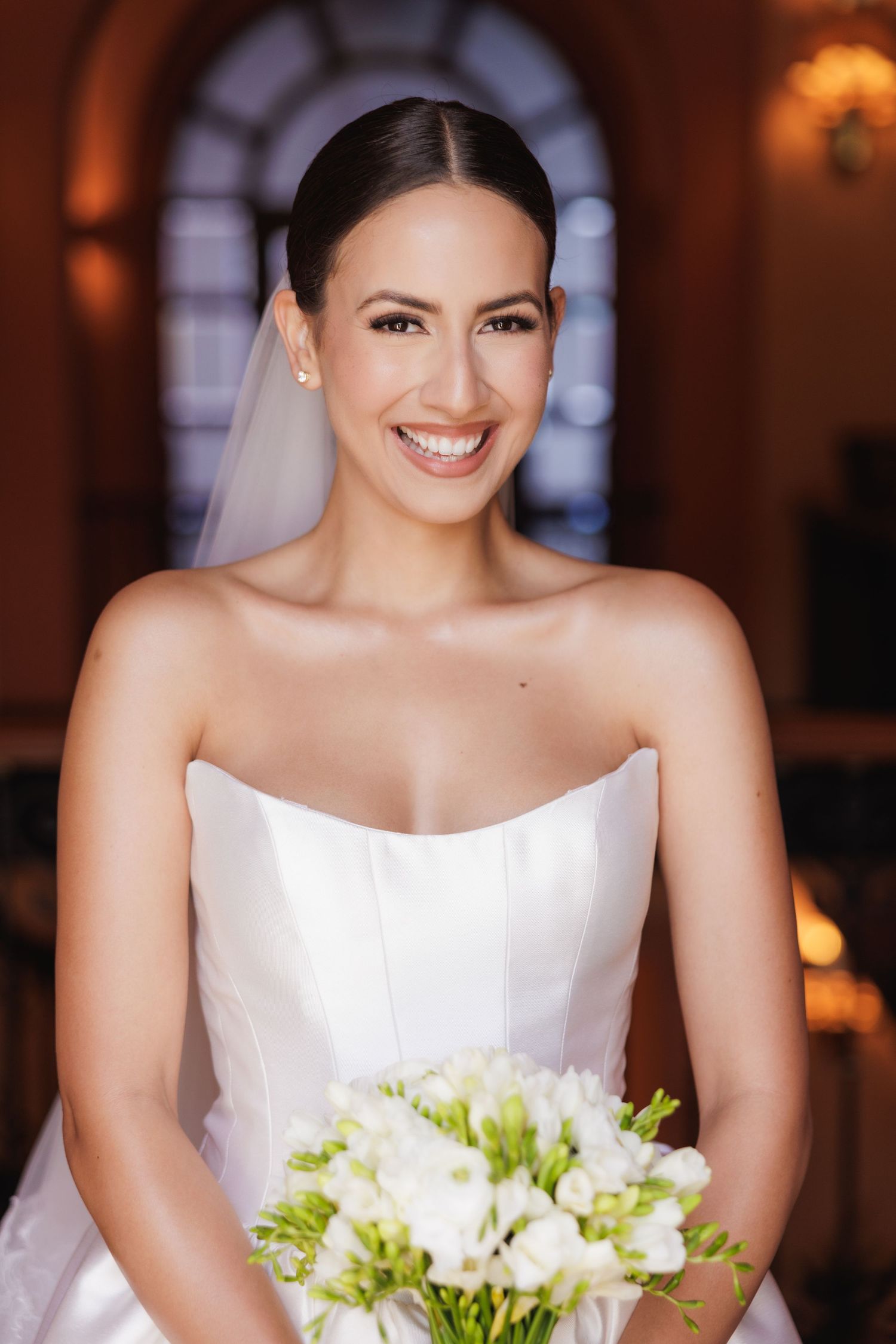 Elegant bride in strapless white gown holds a bouquet of white flowers in a warmly lit venue.