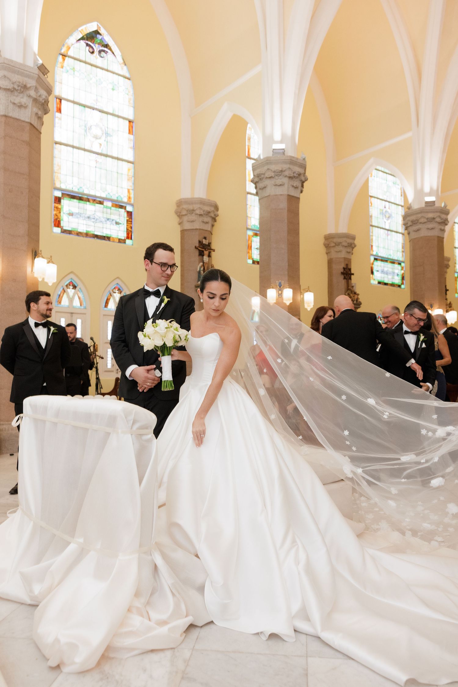 A dramatic wedding dress train flows across a church floor during a ceremony with guests looking on.
