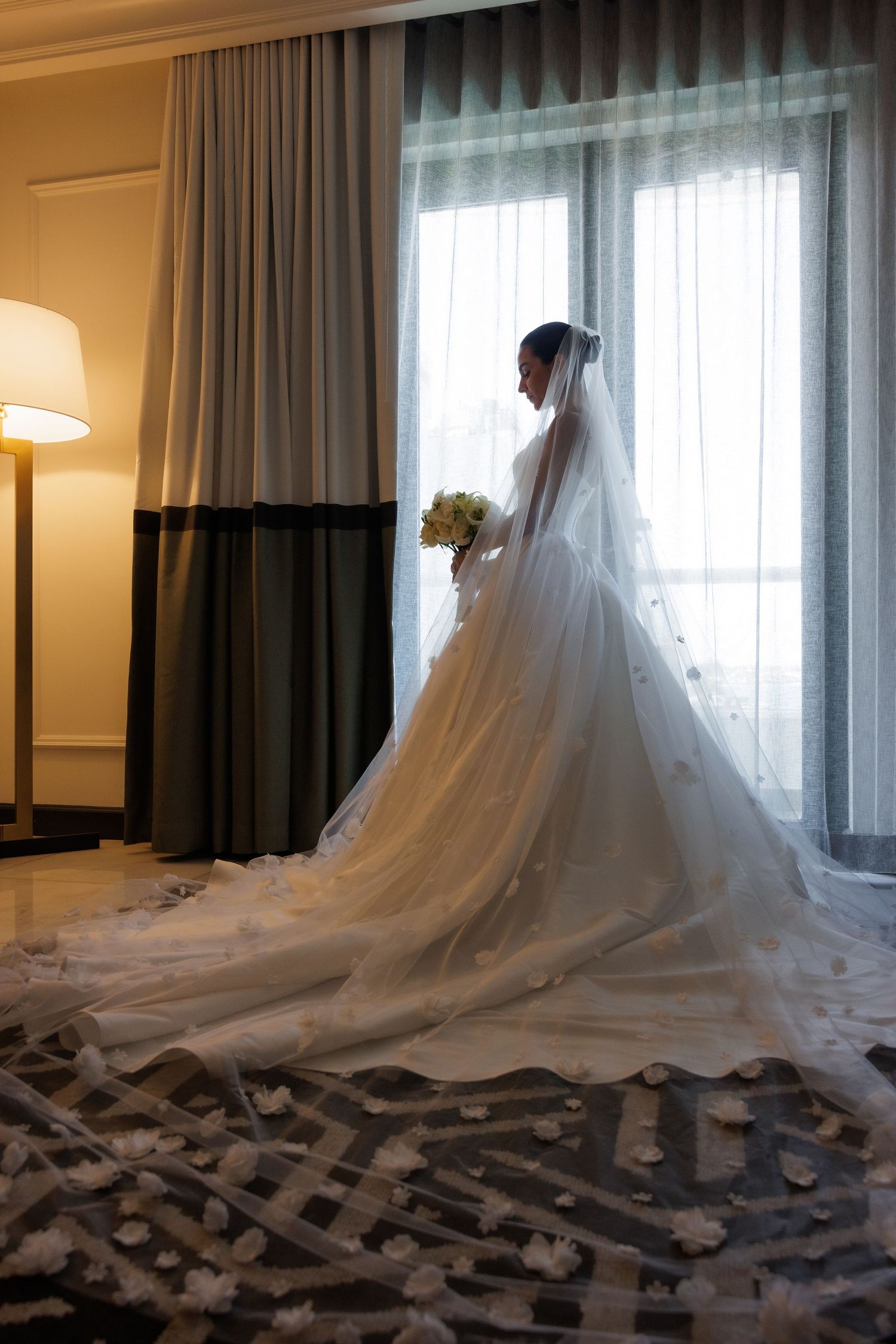 A silhouetted wedding gown with a long veil and train is captured against window light in a hotel room.