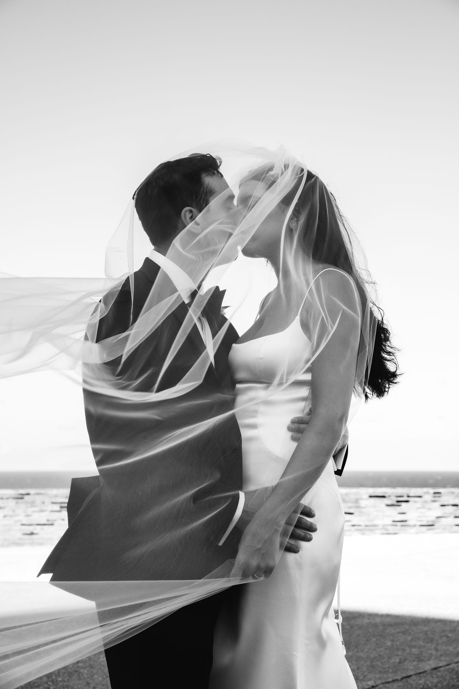 A romantic black and white beach wedding photo of a couple kissing under a flowing veil.