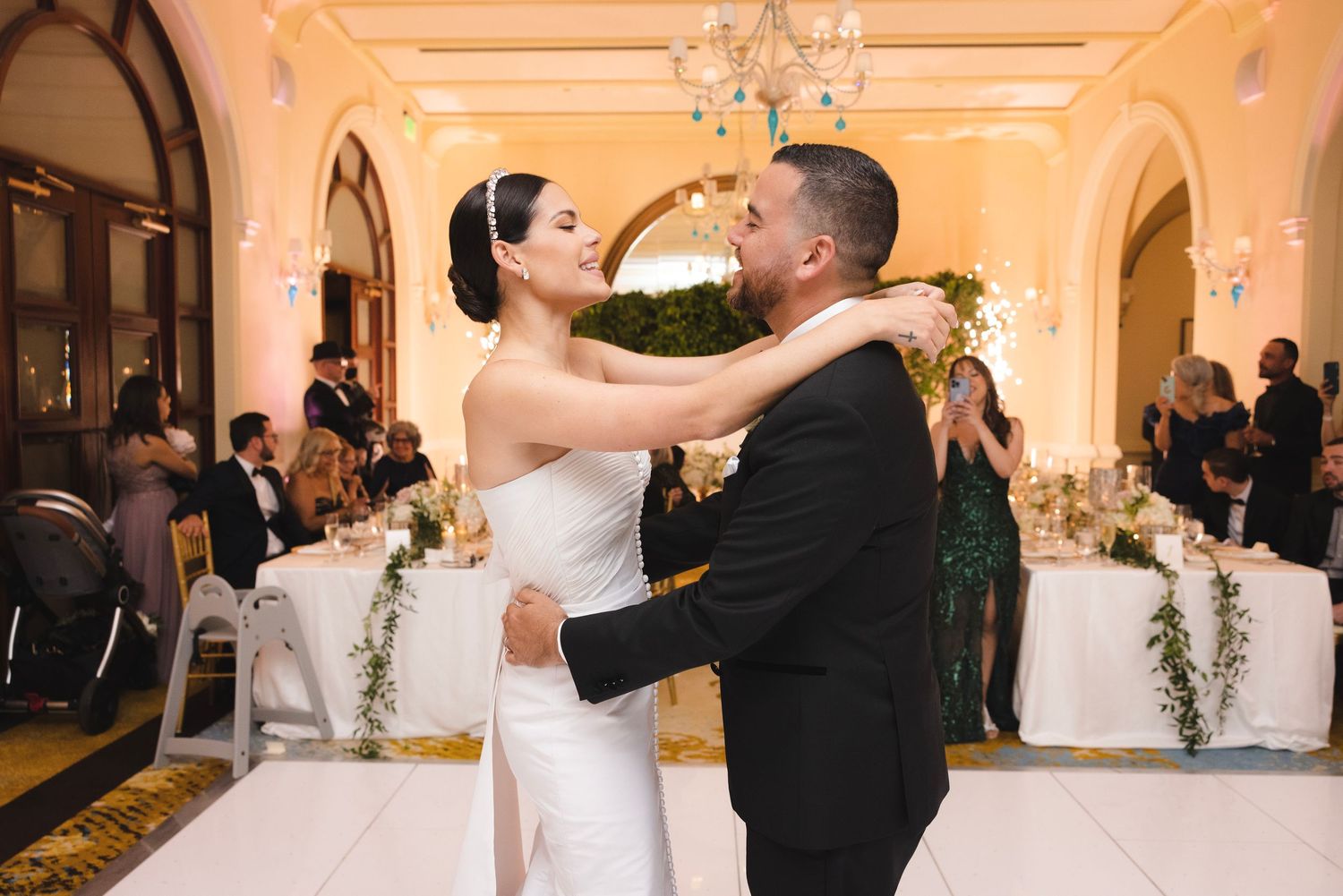 A couple shares their first dance as newlyweds in an elegant ballroom with arched doorways and crystal chandeliers.
