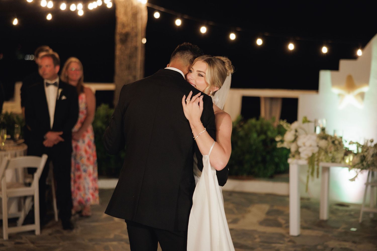 Two people share a romantic dance moment at an outdoor evening reception with string lights overhead.