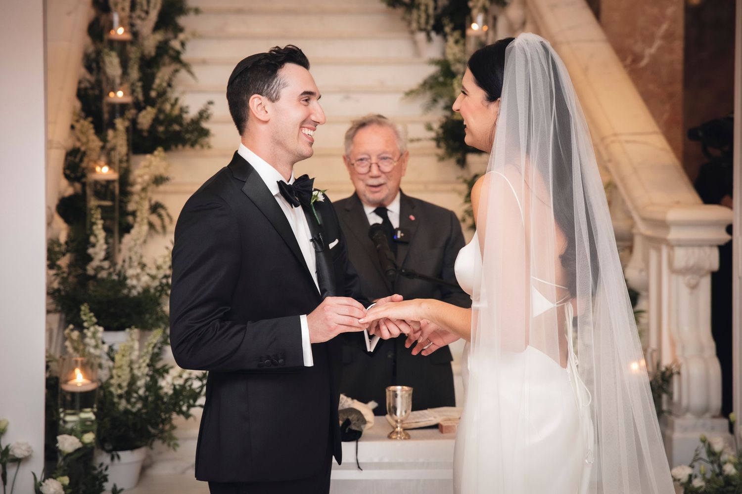 Wedding ceremony captures intimate exchange of rings between couple under decorative floral archway.