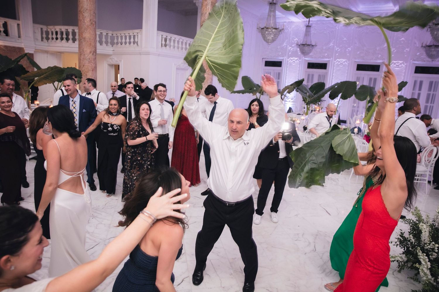 Wedding guests dancing joyfully on a brightly lit dance floor with purple lighting and decorative palm leaves.