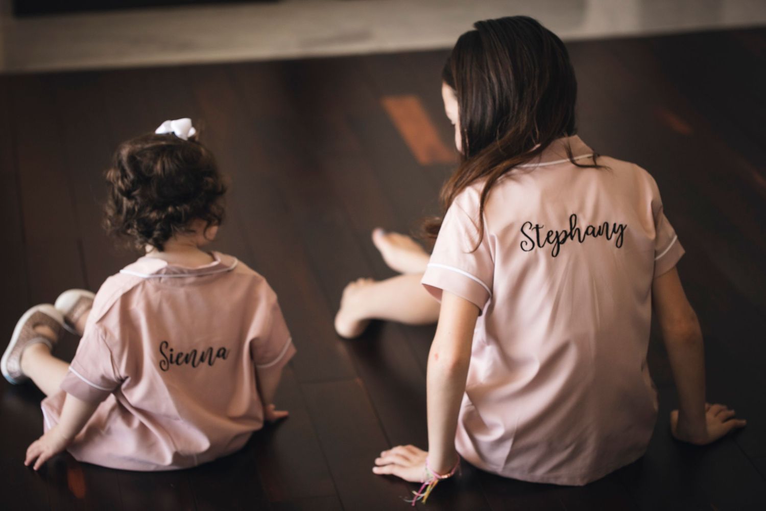 Two people in pink t-shirts with embroidered text sit side by side on a dark floor.