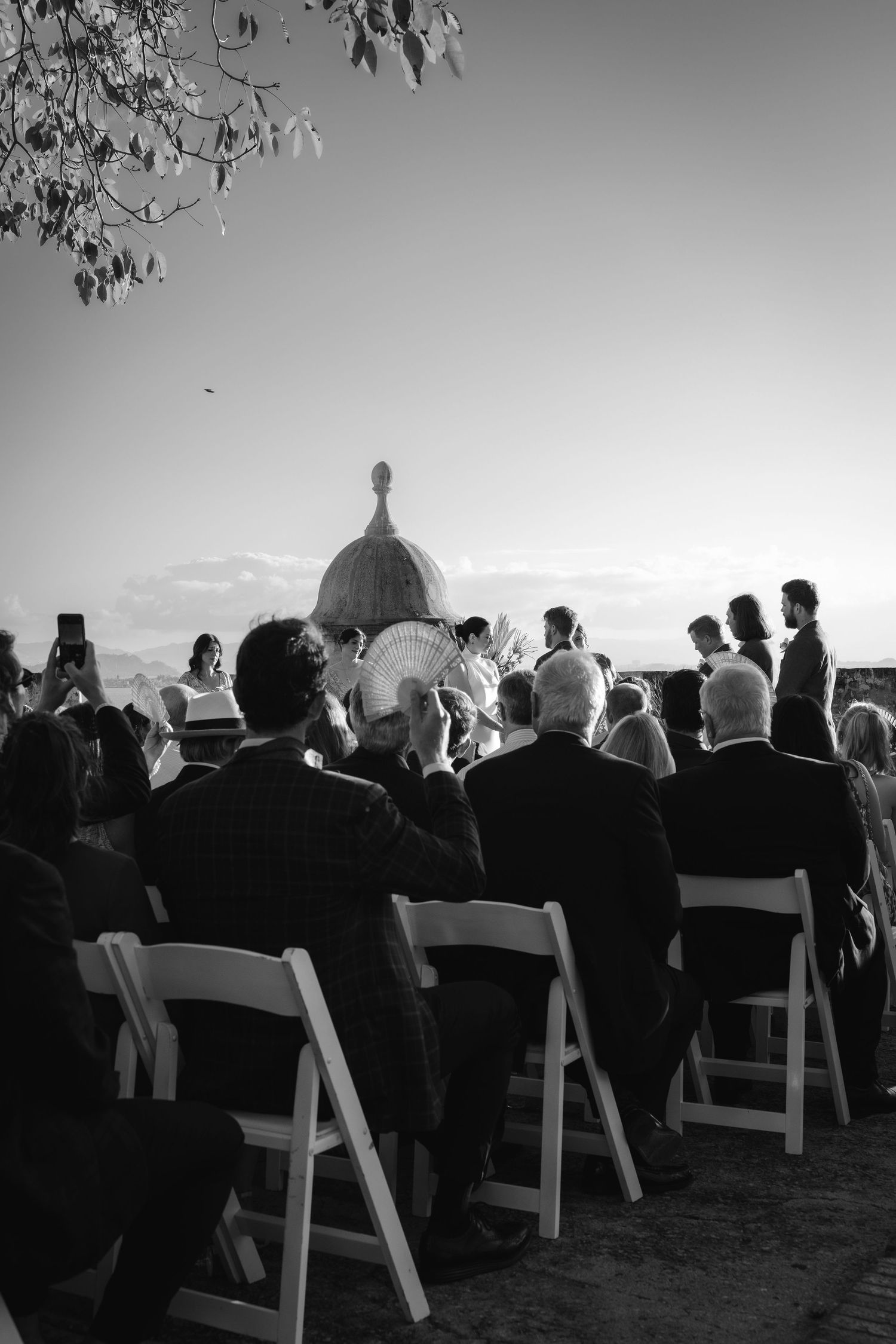 Wedding ceremony attendees seated in white chairs against a sunset backdrop overlooking a scenic view.