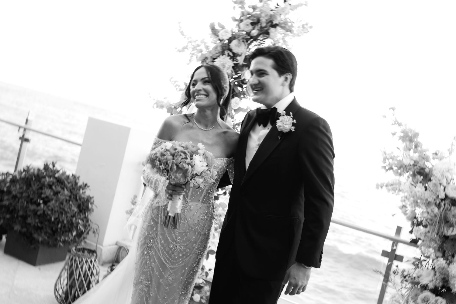 A black and white wedding photo showing a couple smiling outdoors, with the bride holding a floral bouquet.
