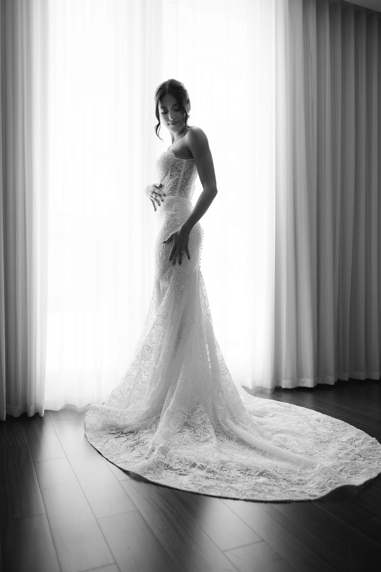 Black and white bridal portrait showing a lace wedding dress with dramatic train silhouetted against window light.