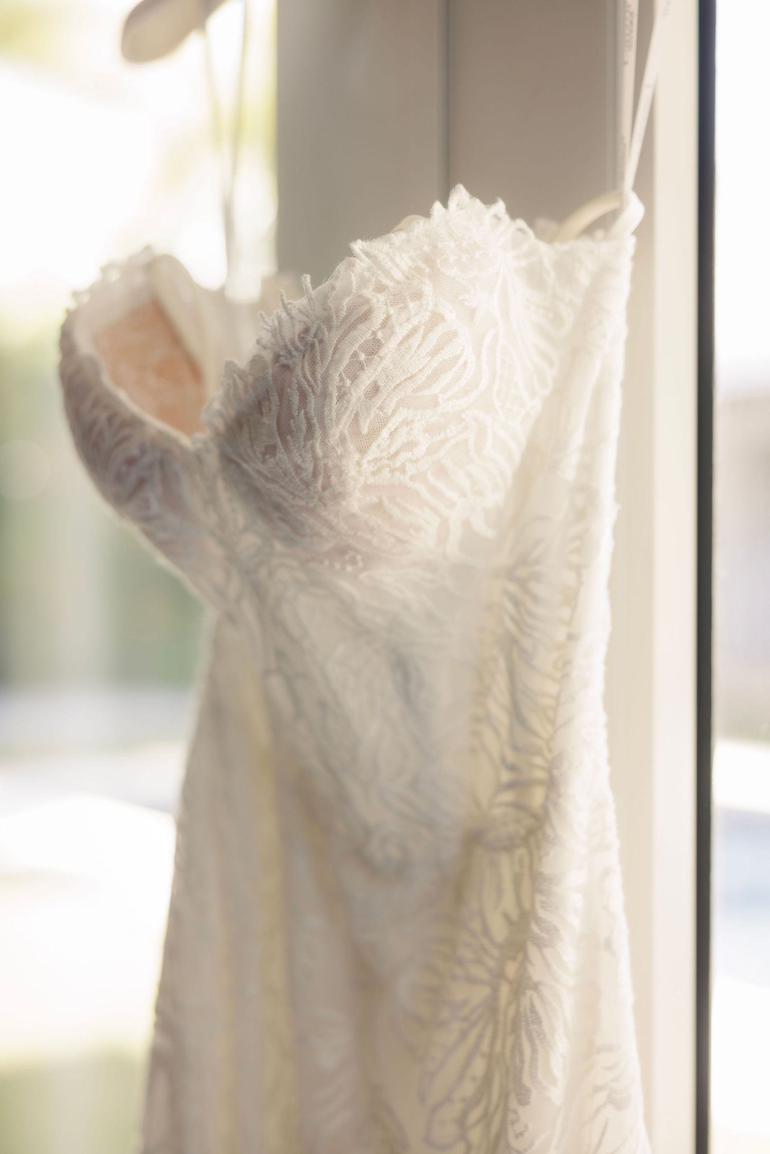 Close-up detail of white lace wedding dress bodice hanging near a sunlit window.