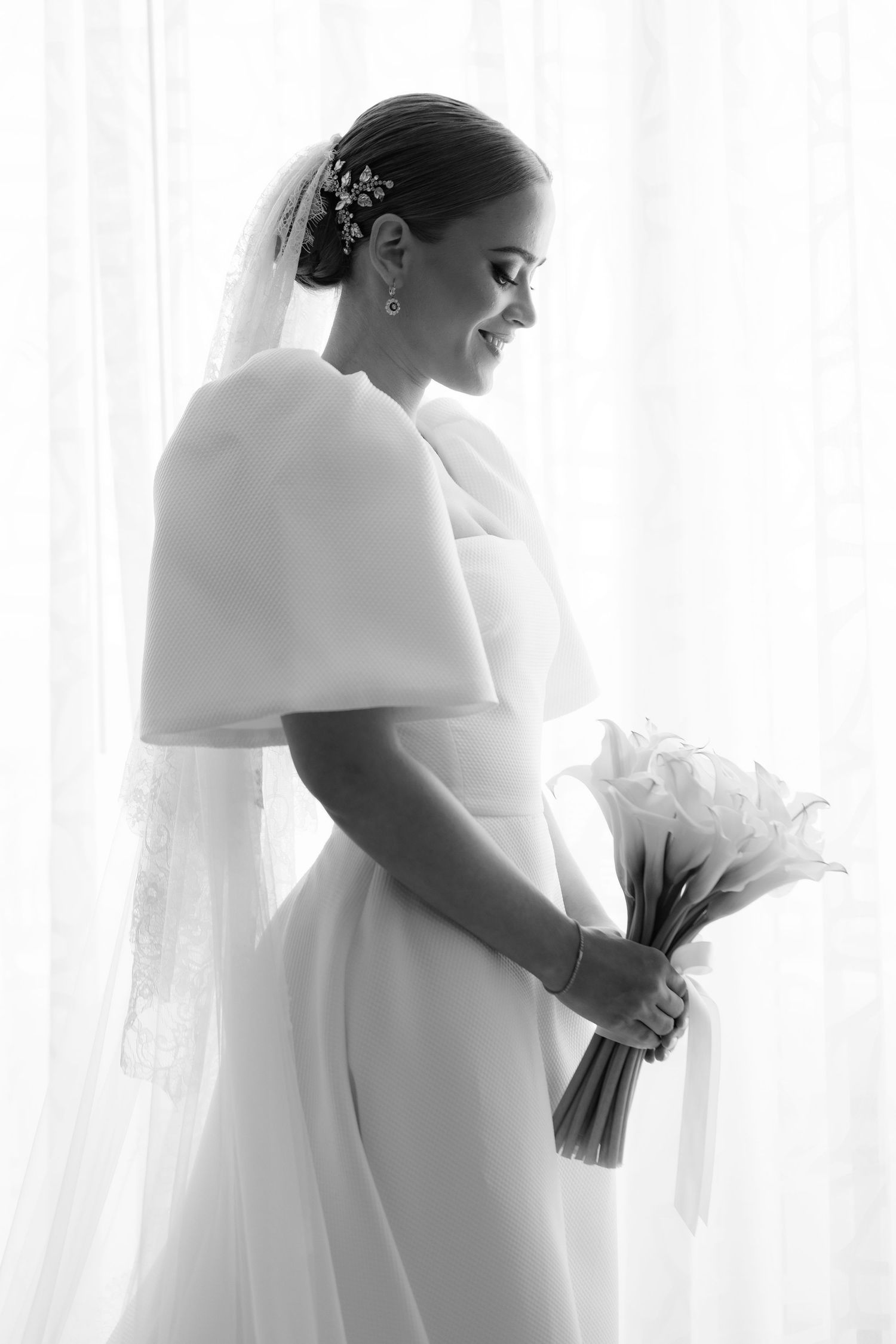 Black and white silhouettes of a bride in traditional gown and veil with a bouquet against white curtained windows.