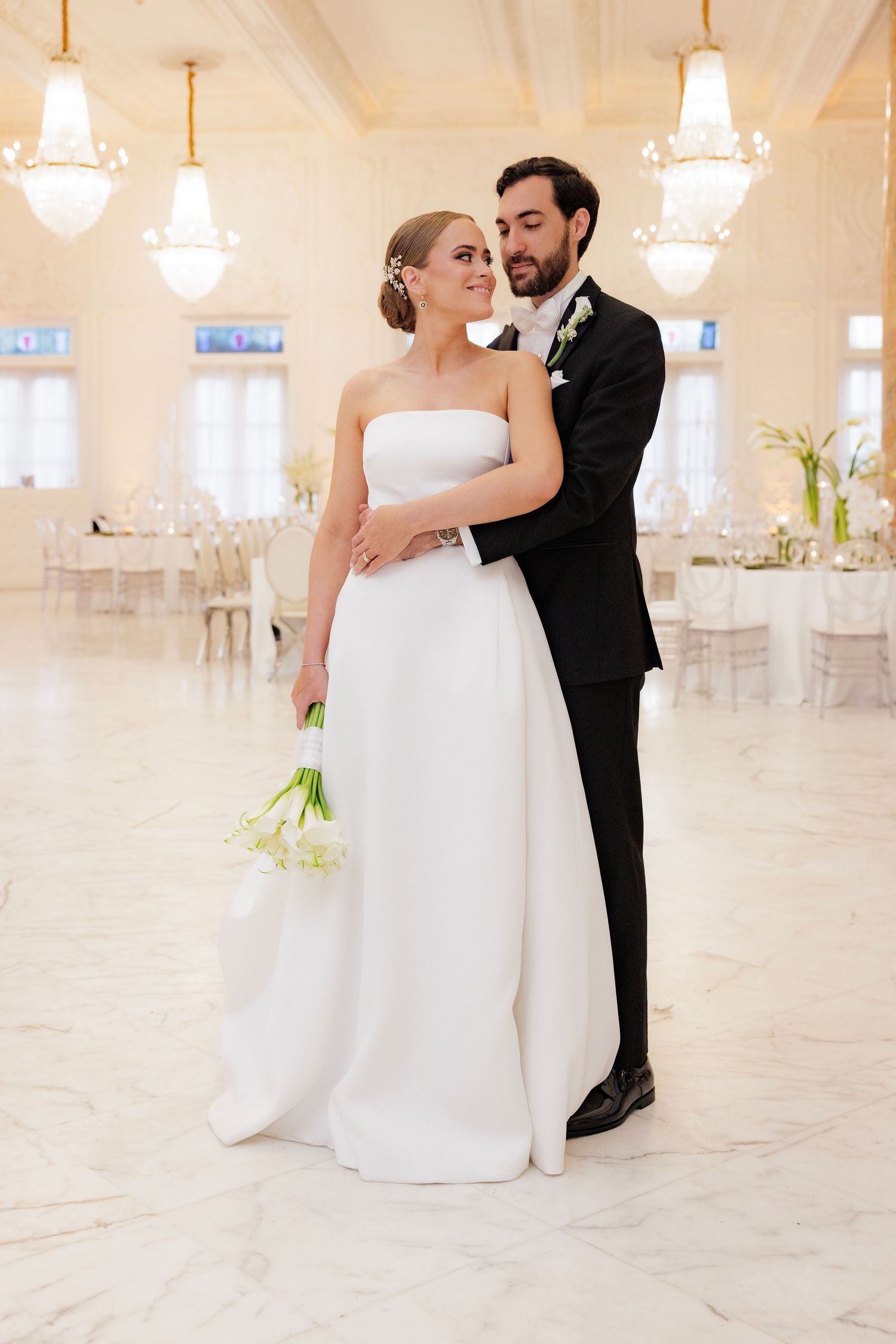 Elegant wedding couple embraces in grand ballroom with crystal chandeliers and white decor.