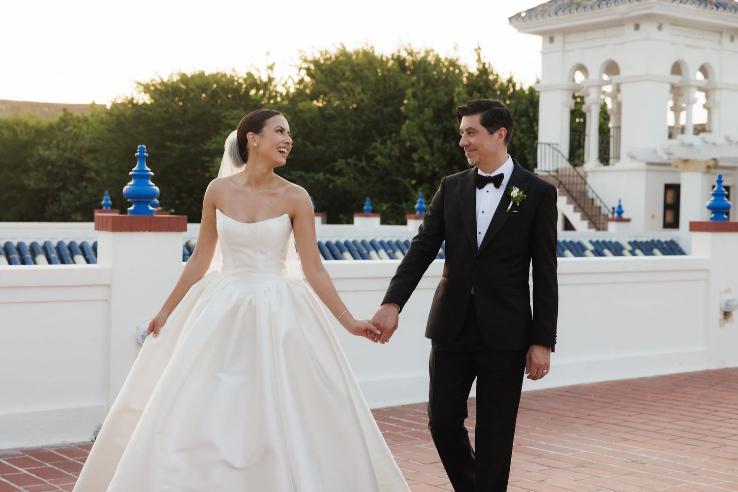 A couple in formal wedding attire strolls together on a white terrace with architectural details and greenery behind them.