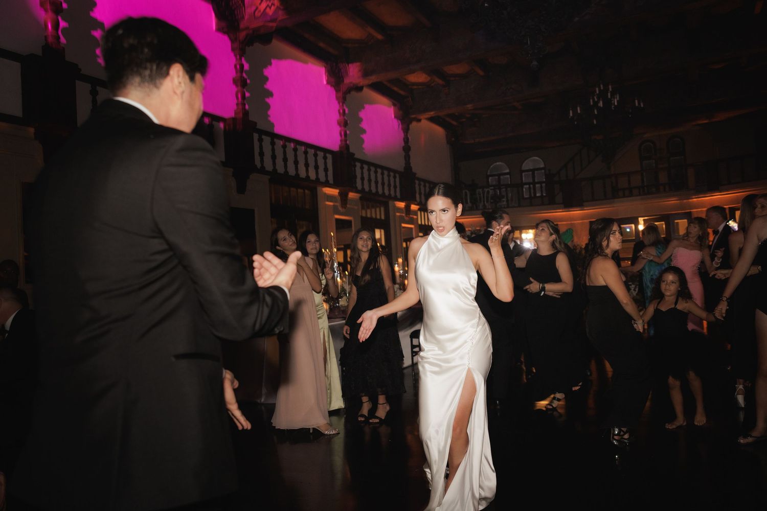 People dancing and celebrating at a wedding reception in a dimly lit venue with pink lighting accents.