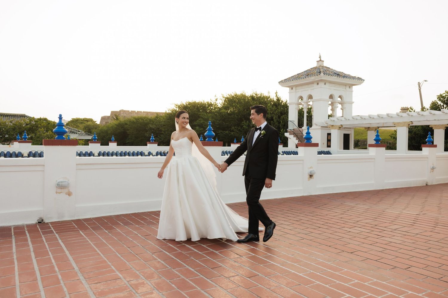 A couple in wedding attire walks along a white tiled outdoor terrace overlooking a scenic coastal view.