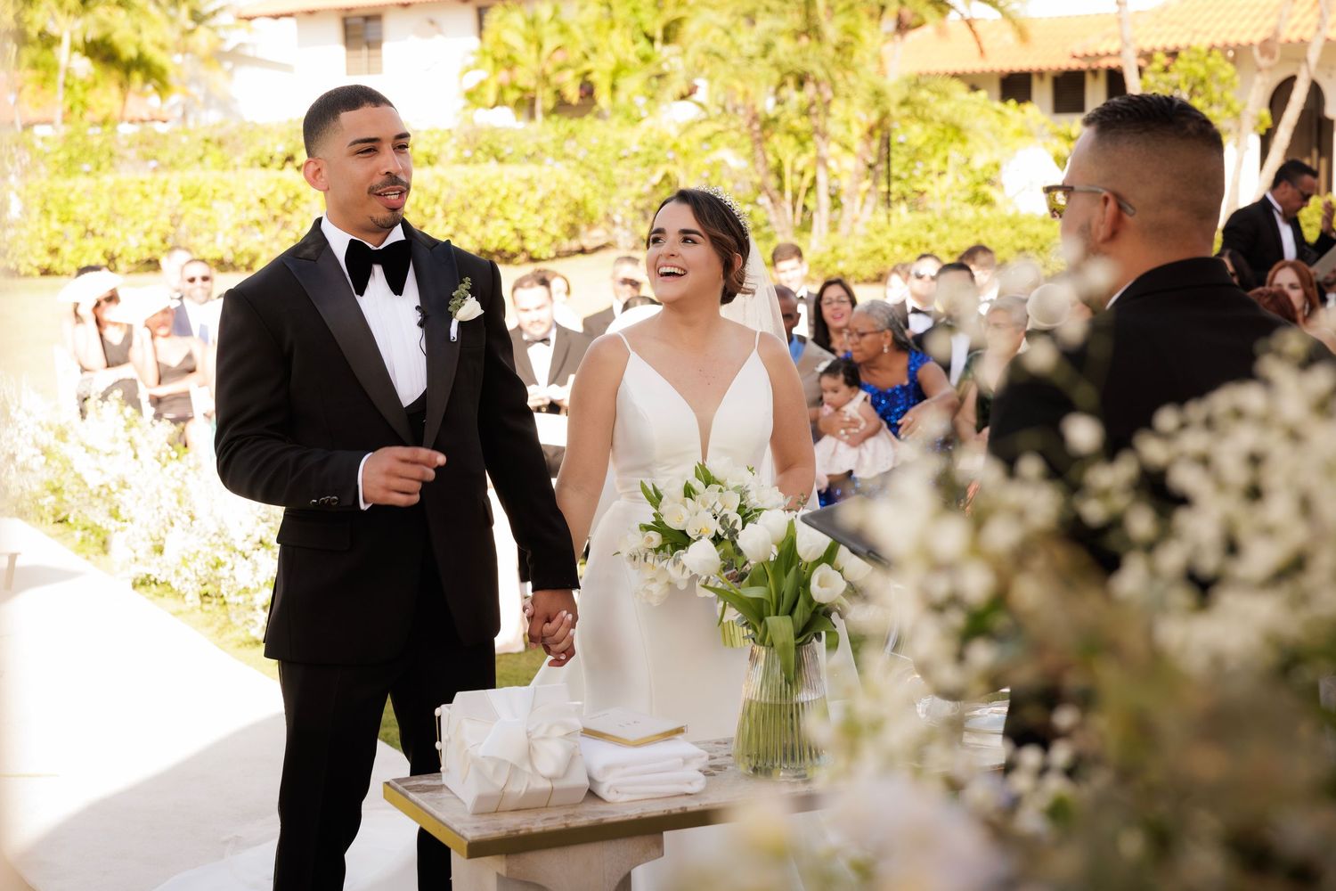 A couple celebrates at their outdoor wedding ceremony under a sunlit pergola with white floral decorations.