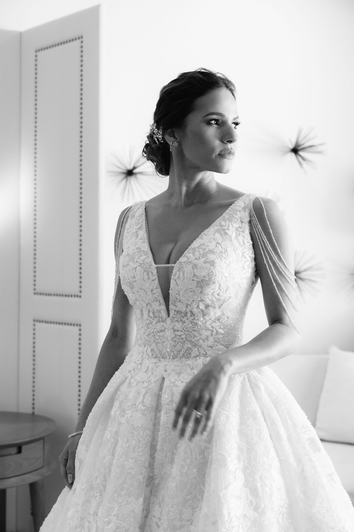 Elegant bride in white lace V-neck ballgown poses gracefully by window in black and white photography series.