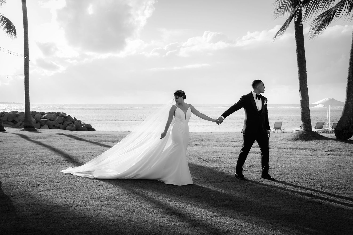 A romantic black and white beach wedding photo with couple holding hands under palm trees by the ocean.