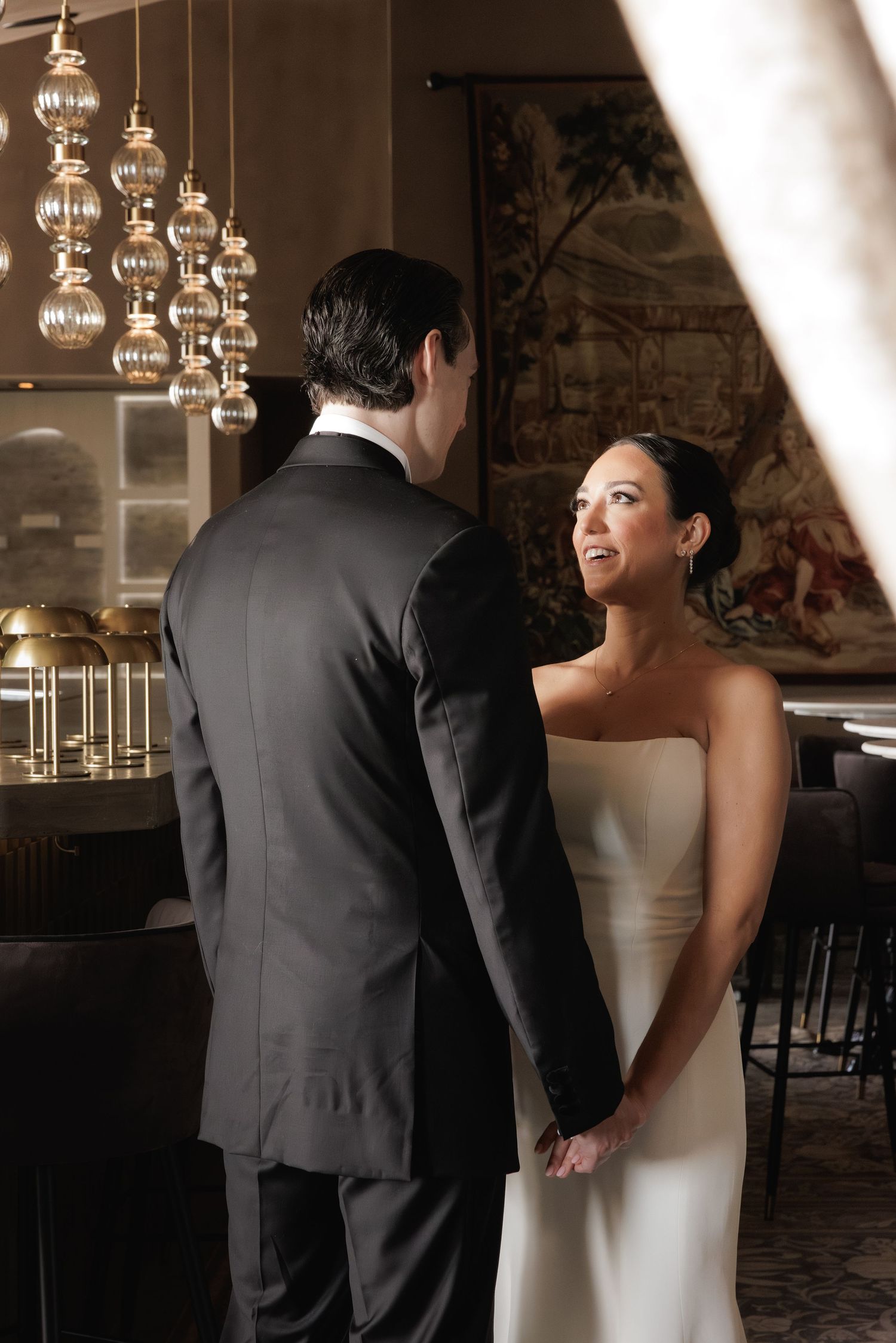 A couple shares an intimate moment in formal attire under elegant glass pendant lights in an upscale venue.