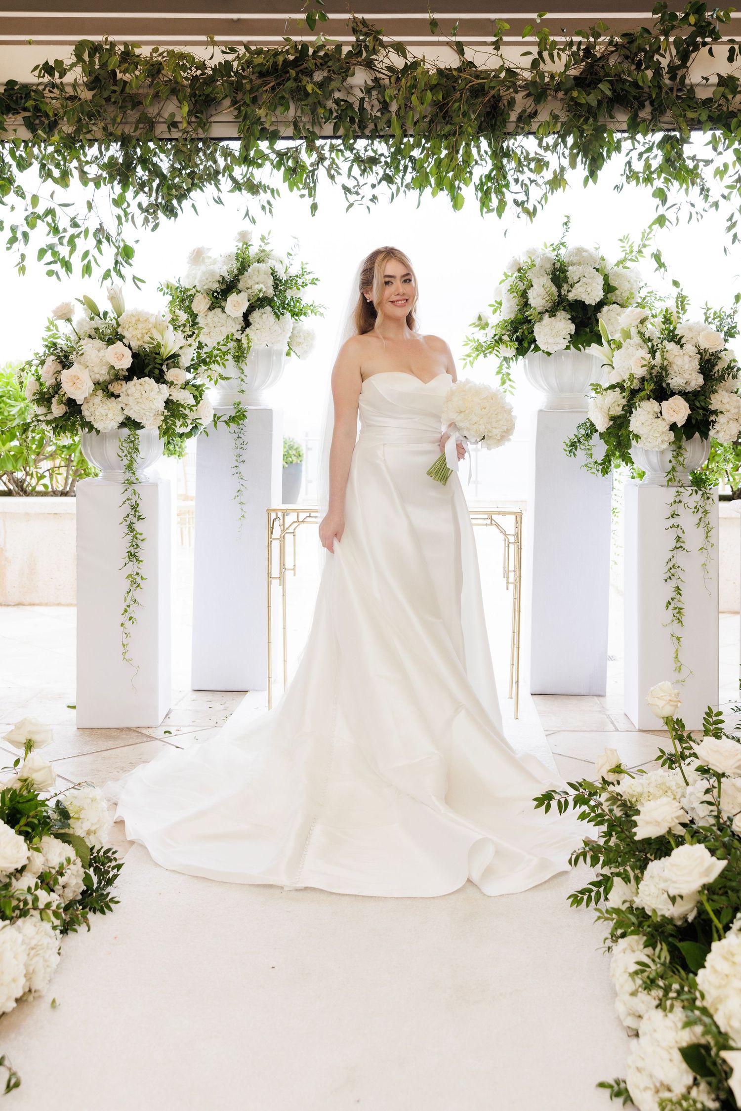 Bride in classic white gown under floral arch with cascading white flowers and greenery.