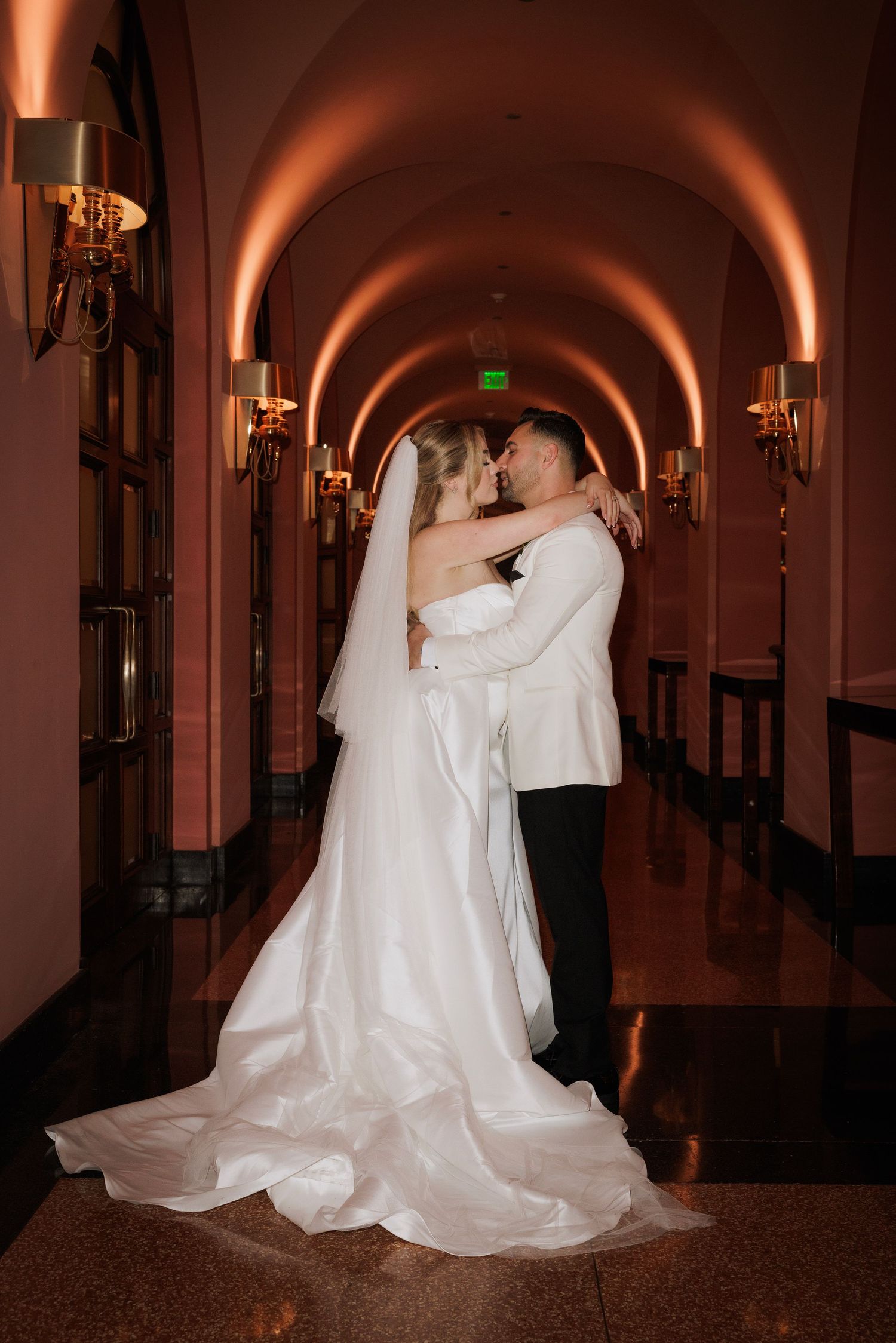 A couple in formal wedding attire dances in a dimly lit hallway with arched ceilings and elegant wall sconces.