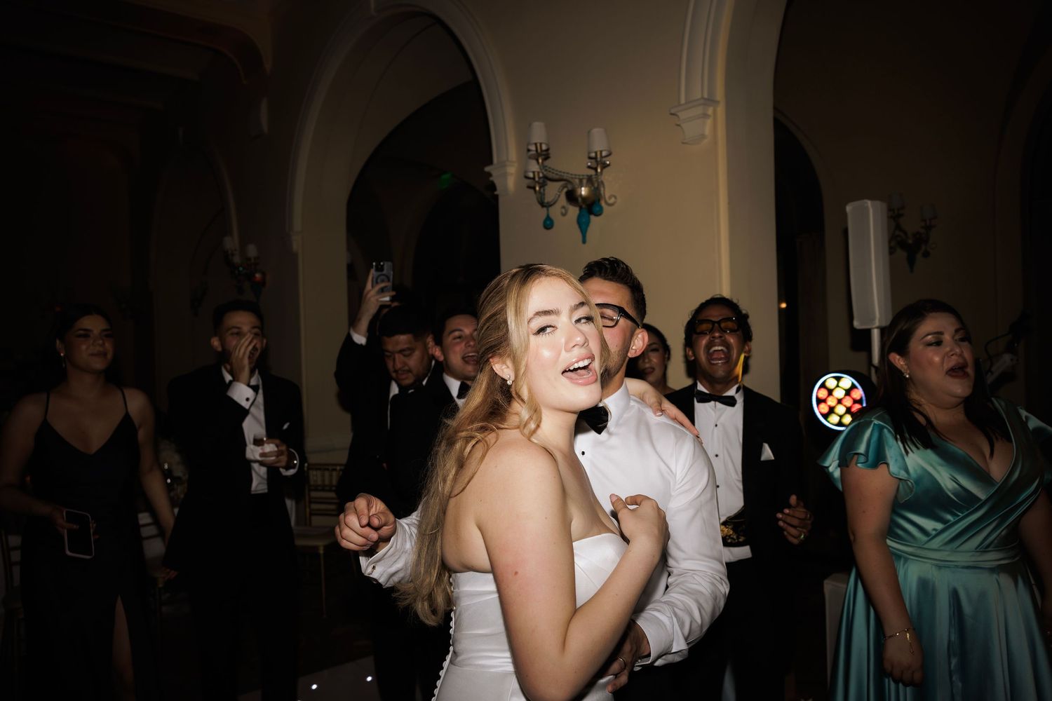 Wedding reception dance party with guests celebrating in formal attire under dimmed lighting.