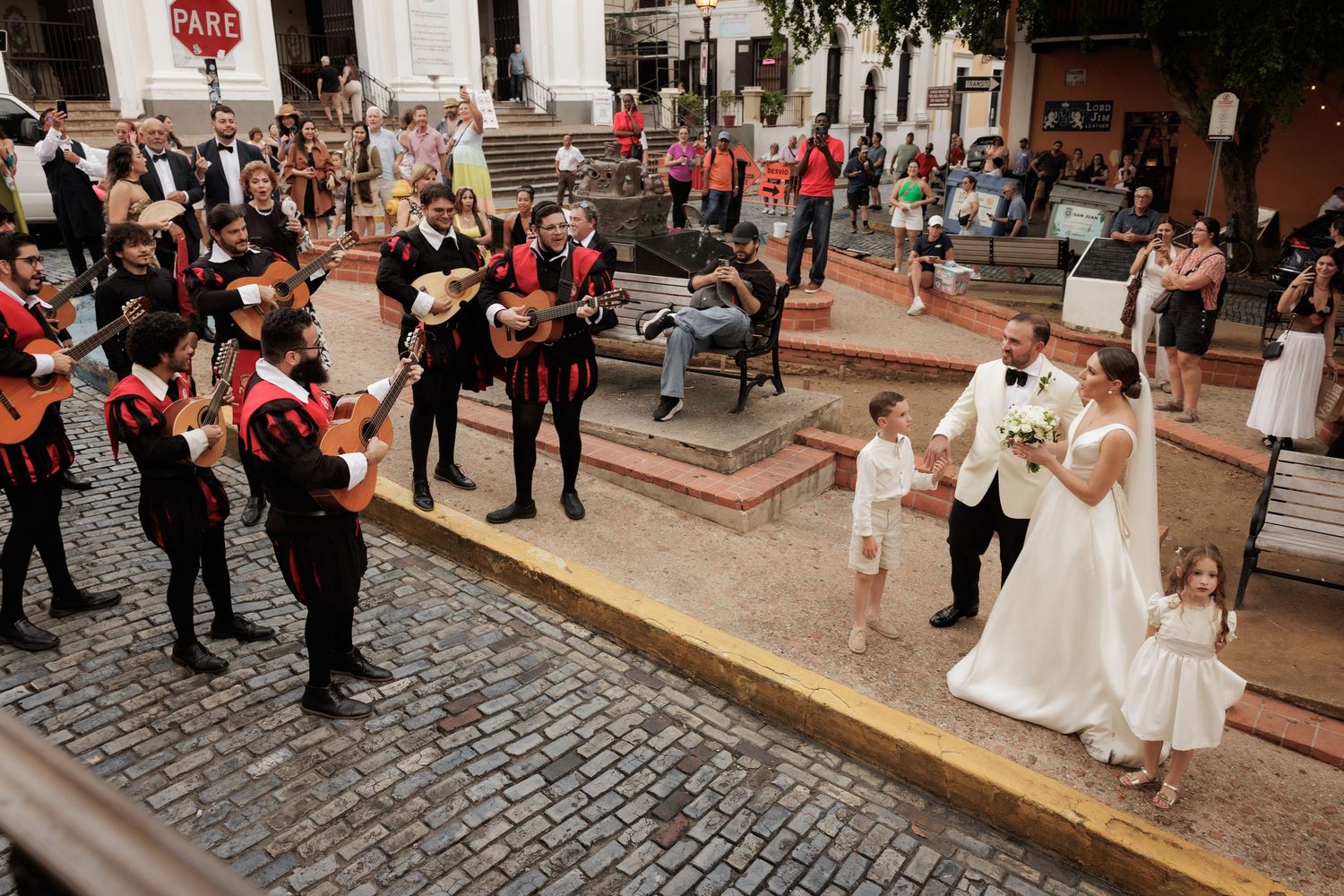 Wedding party members in red and black hold a flowing red fabric on a cobblestone street during a celebration.