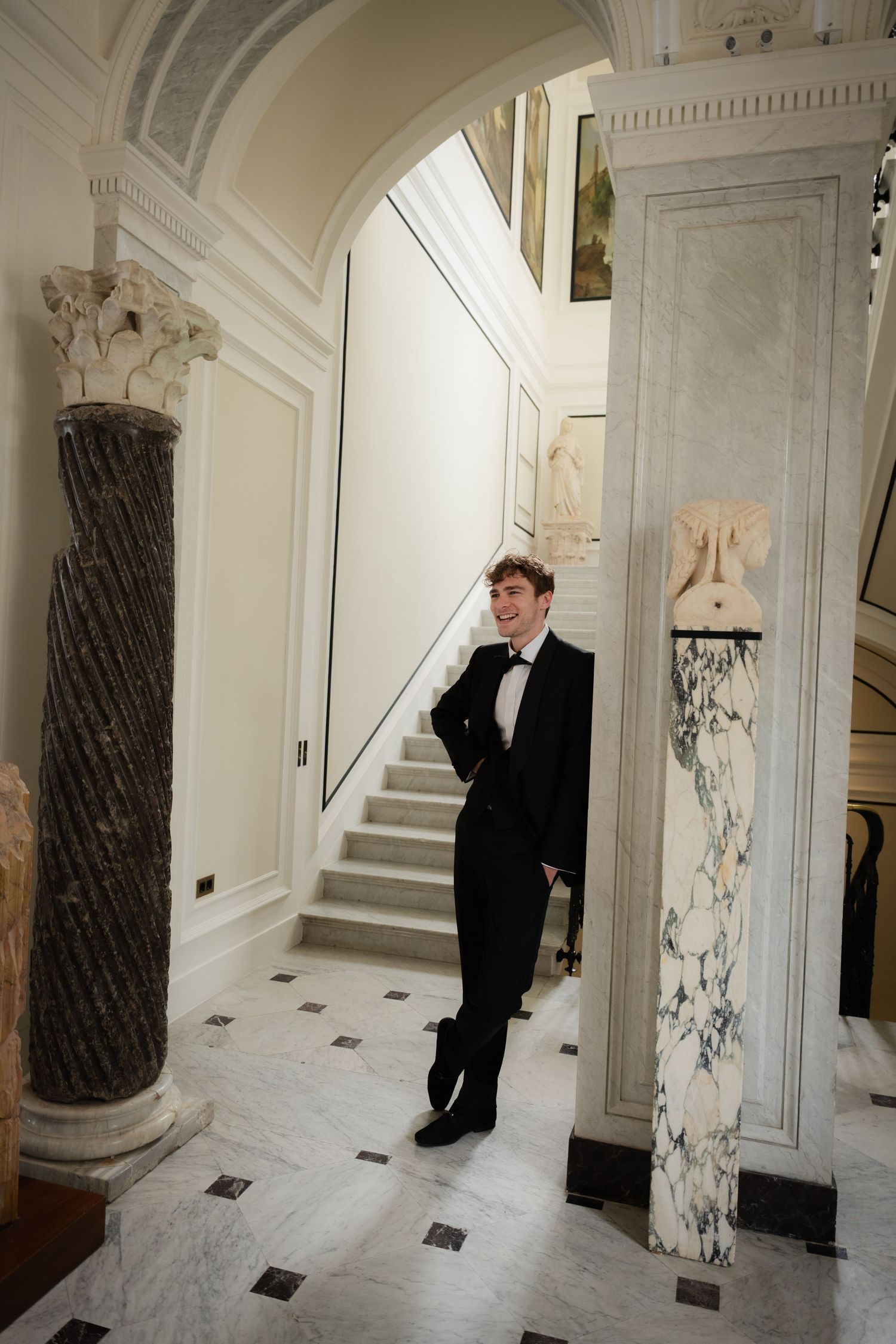 Young man in black tuxedo and bow tie standing between marble columns in an elegant classical interior.