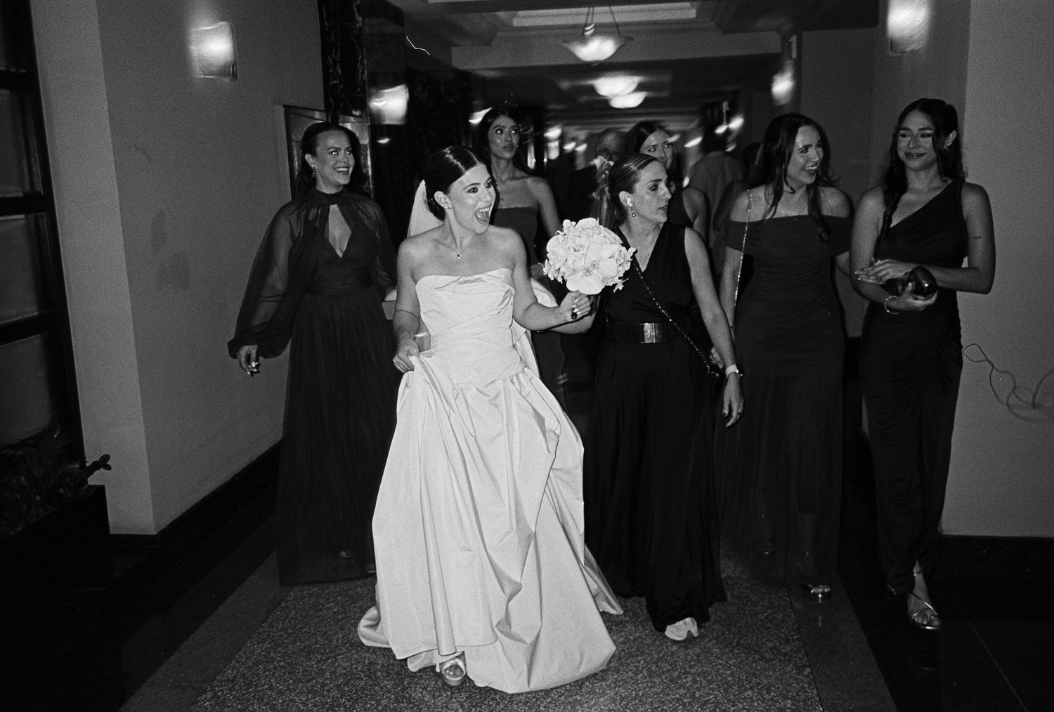 Bride in white gown walks with bridesmaids in black dresses through a hallway, black and white wedding photo.