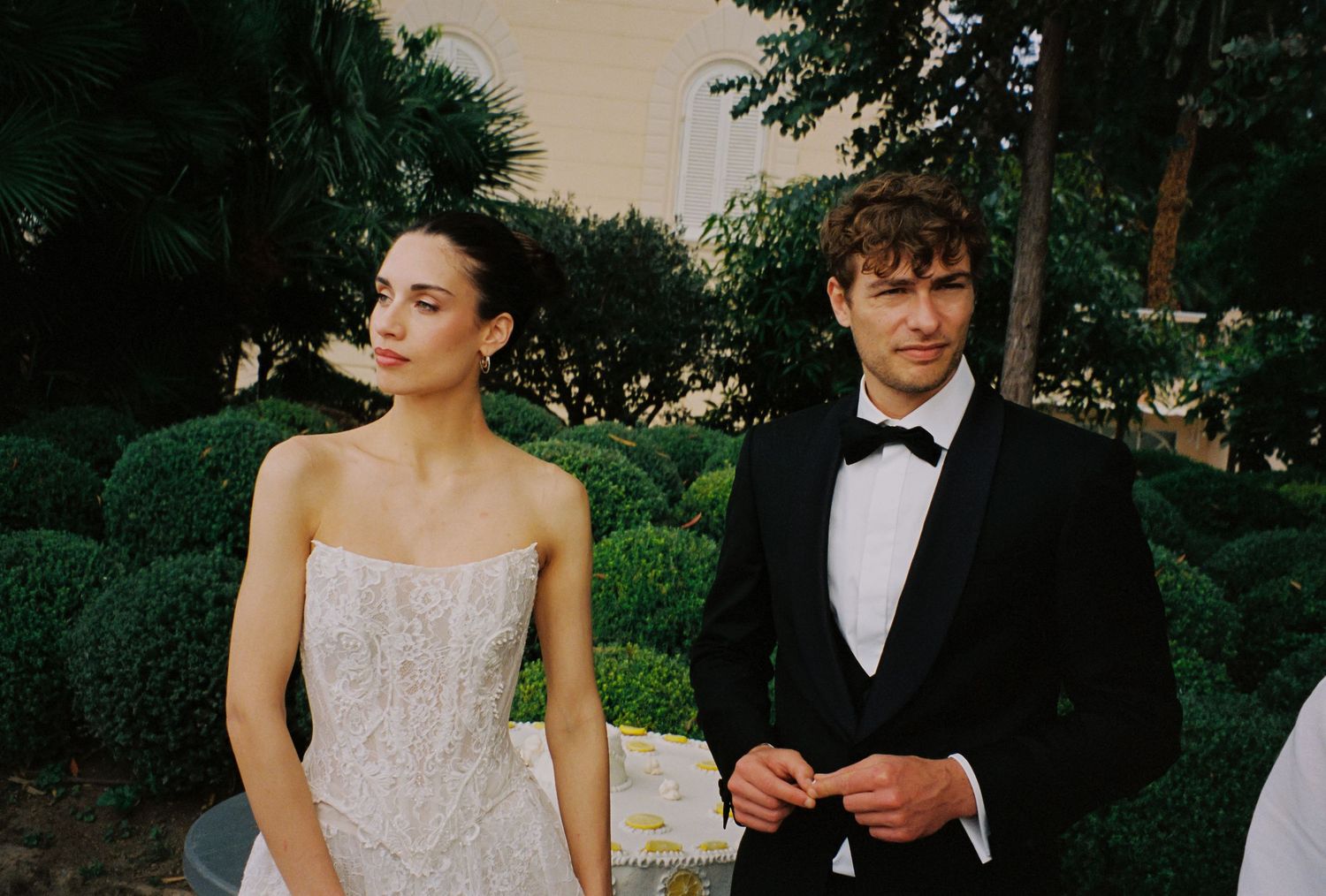 Elegant couple at a wedding, woman in strapless lace gown and man in black tuxedo with bow tie outdoors.