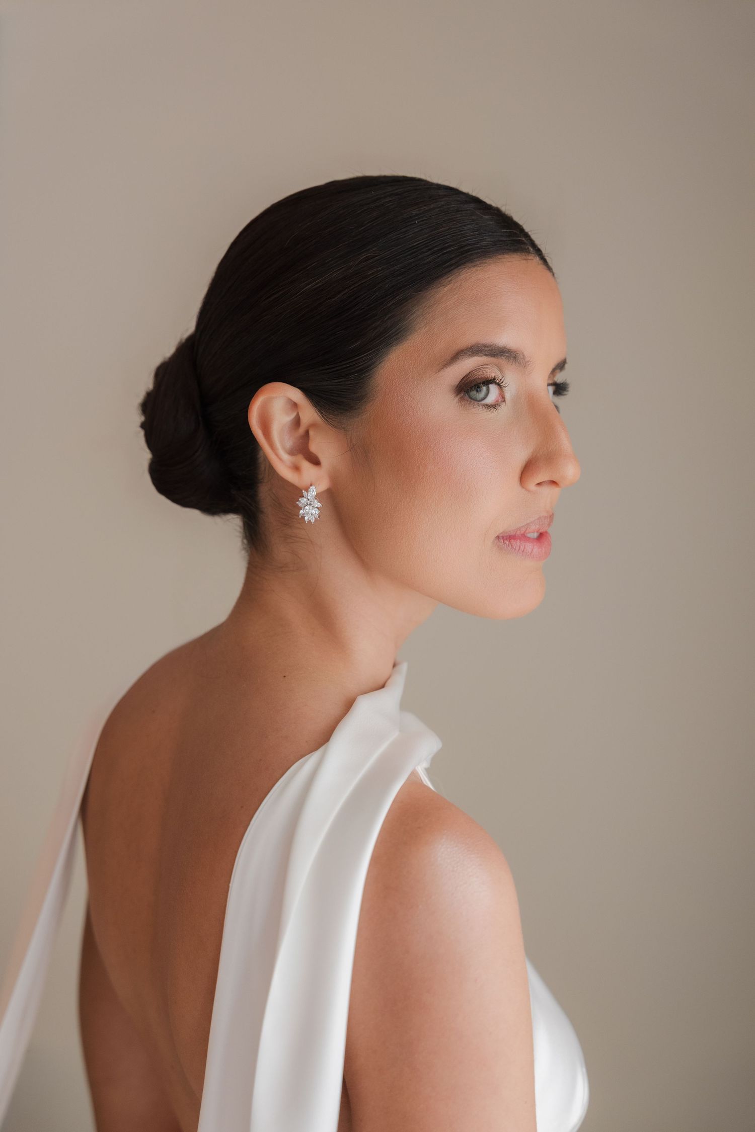 Elegant bride in backless white gown with crystal drop earrings and sleek updo hairstyle, side profile view.