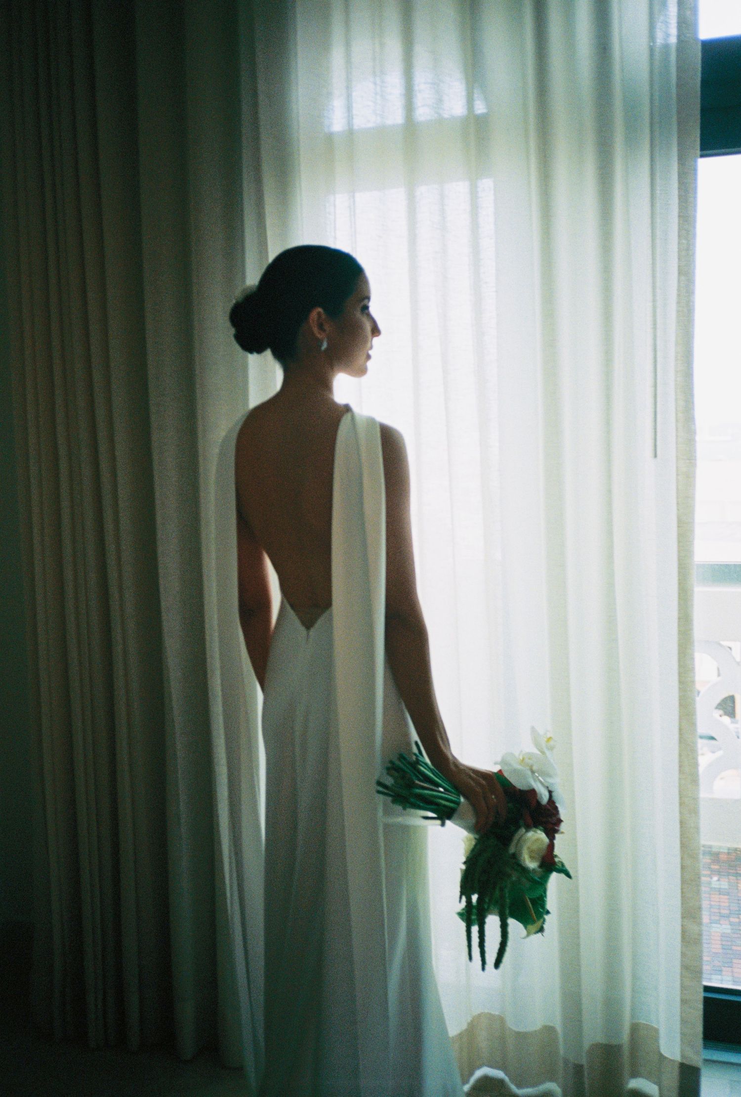 Elegant bride in backless white gown holding bouquet, standing by sheer curtains in soft natural light.