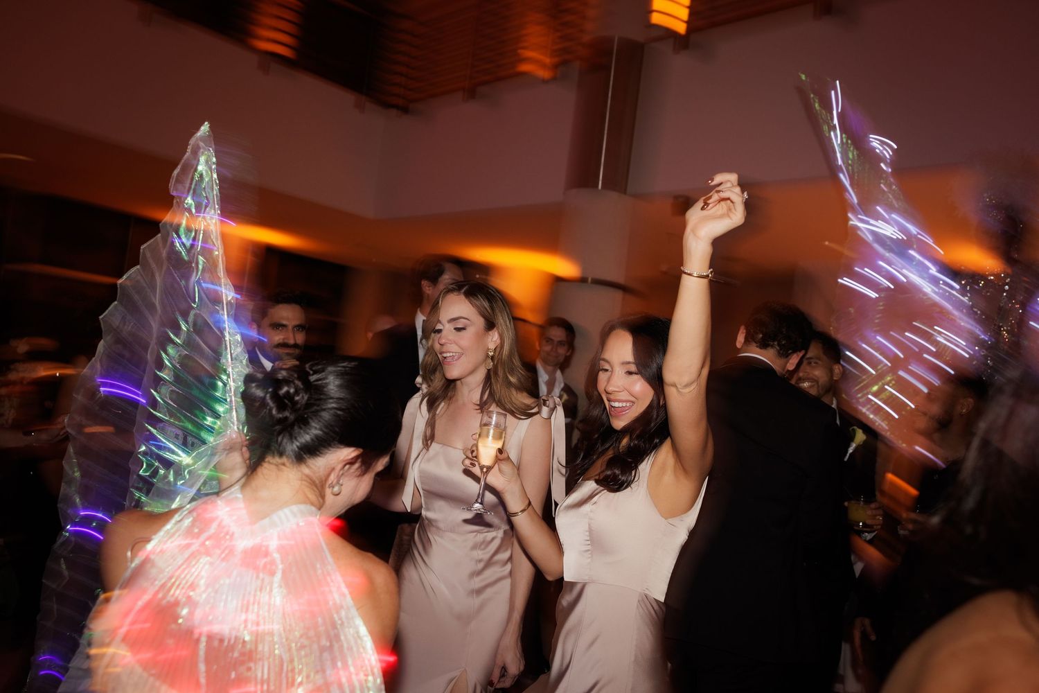 People dancing and celebrating at a party with colorful LED light wands creating vibrant streaks in the dimly lit room.
